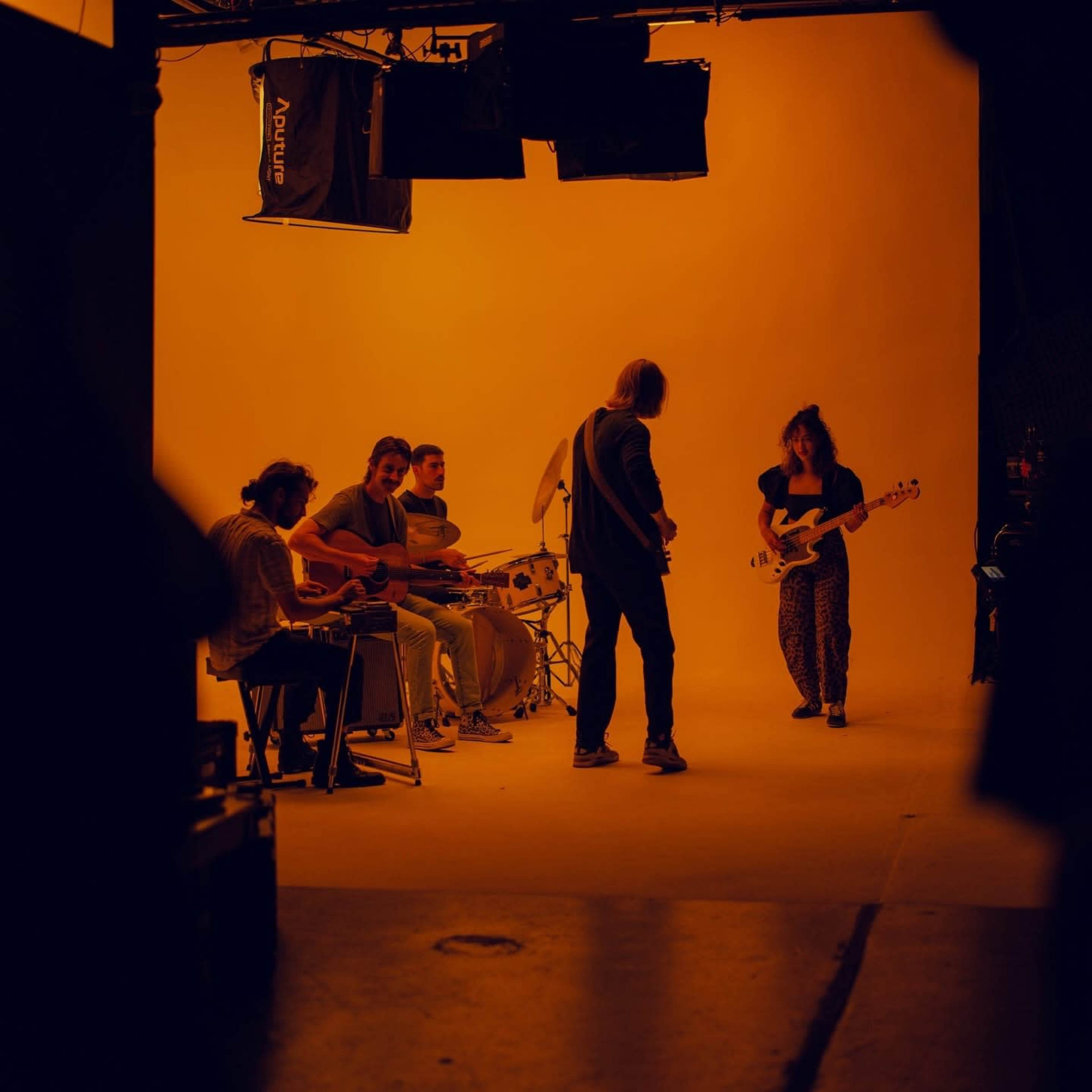 A band performs on a stage with a bright orange backdrop, featuring instruments like guitars and drums.