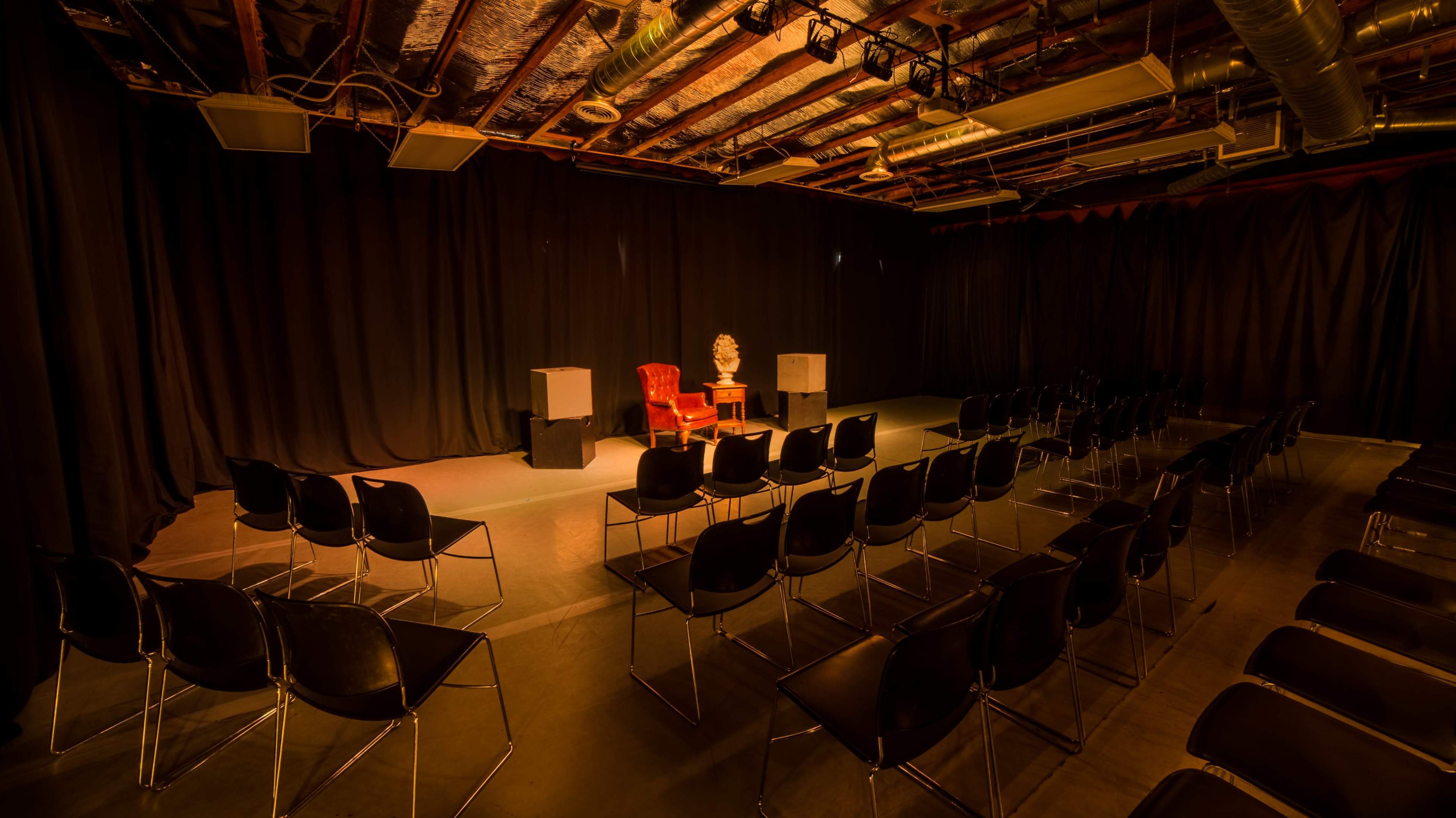 The image shows a dimly lit theater space with a pair of red chairs facing a small table, surrounded by rows of black chairs.