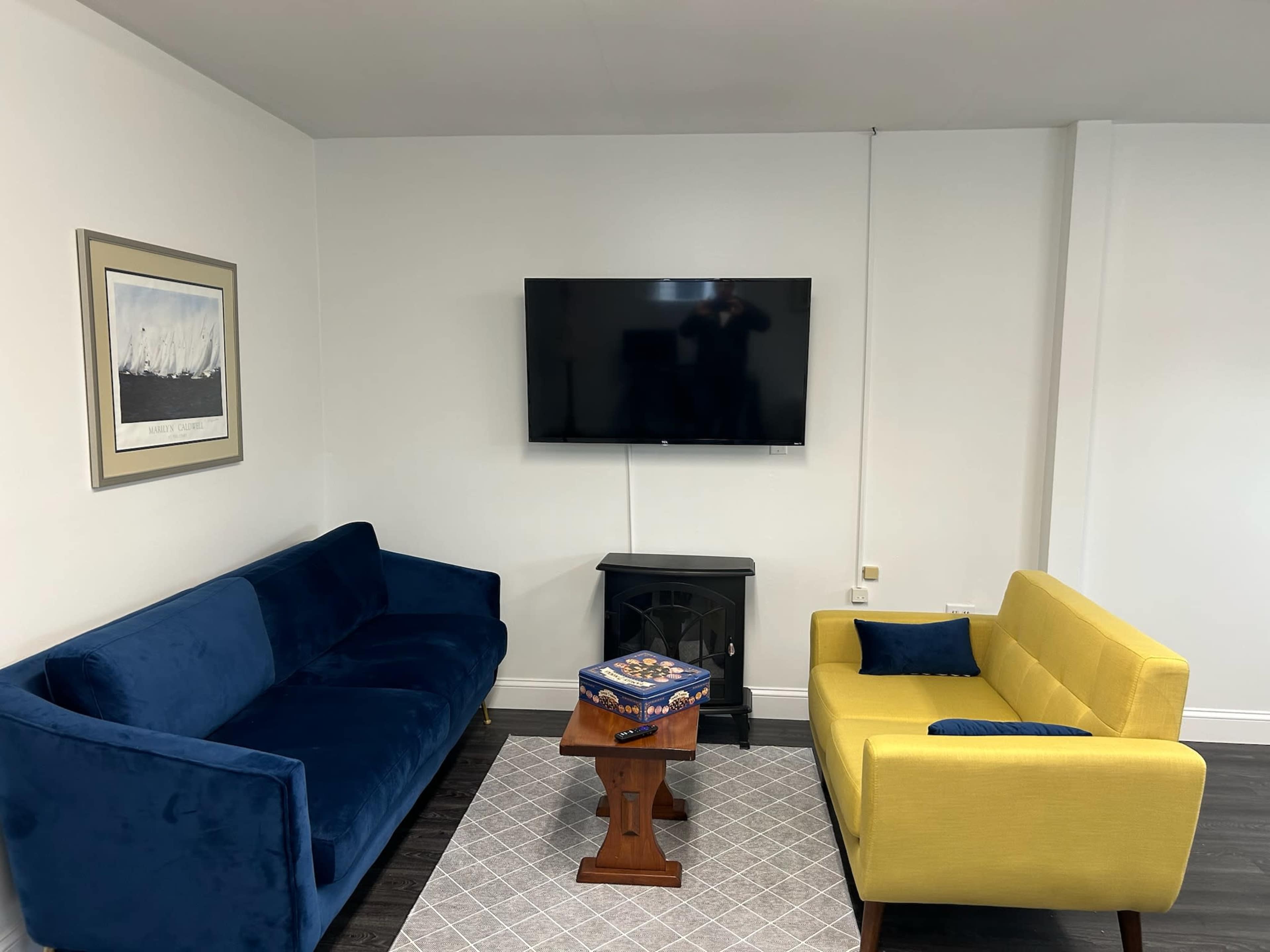 The image shows a living room with a blue sofa and a yellow sofa facing a wall-mounted television, along with a small wooden table and a decorative rug on the floor.