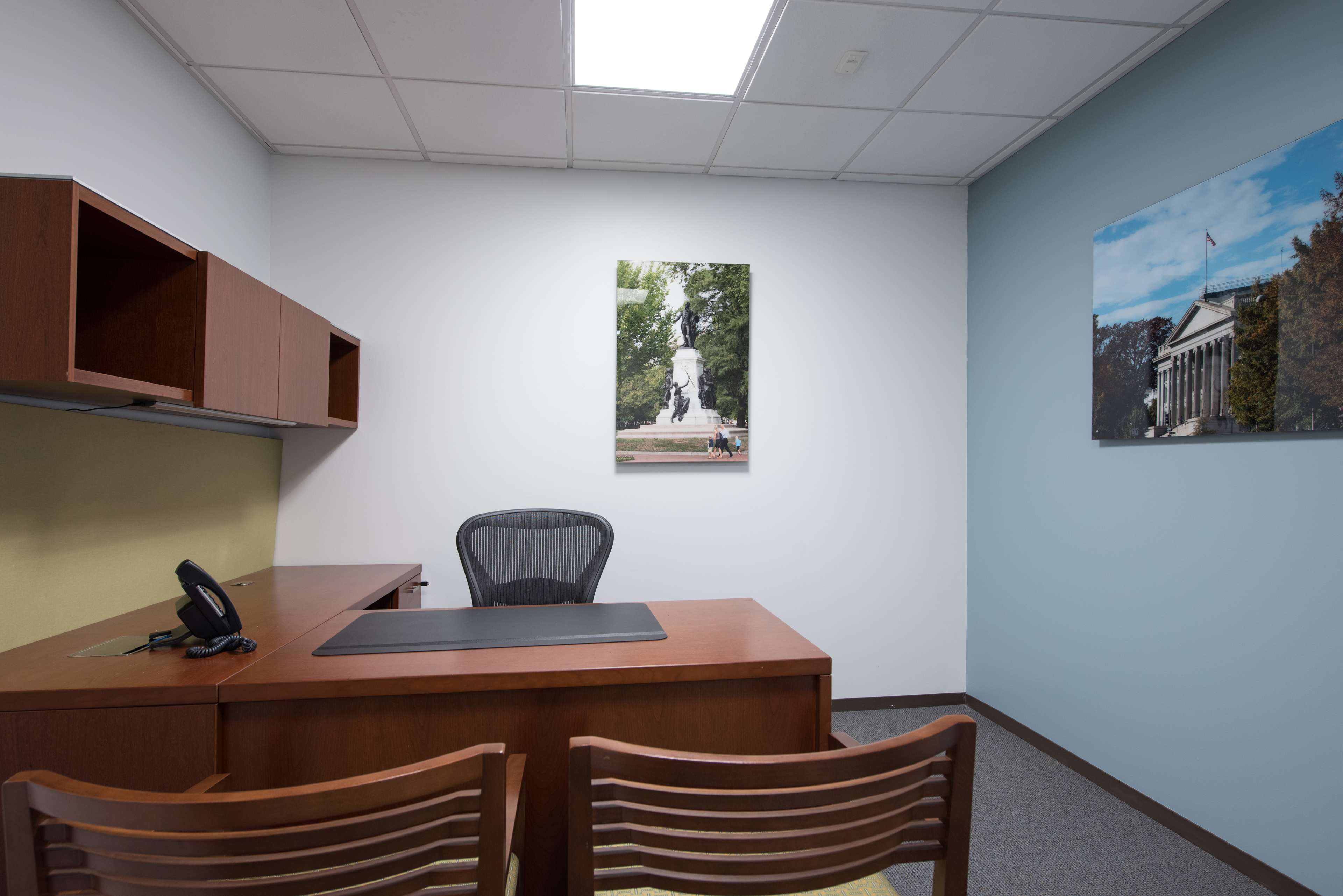 A small office with a wooden desk and chair, a phone on the desk, and two wooden chairs facing it, with framed photographs of a statue and a building on the walls.