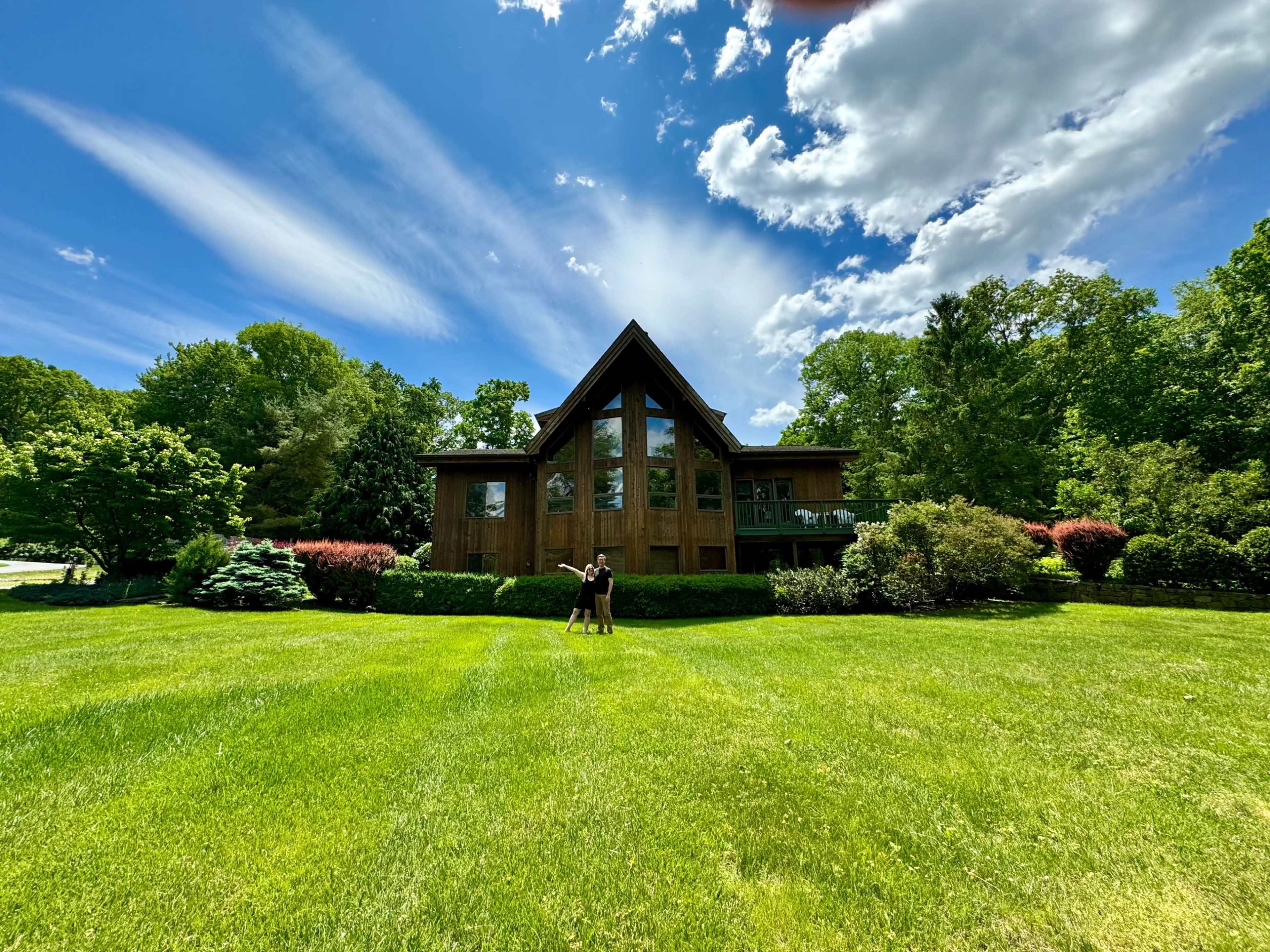 A large wooden house with a peaked roof sits in a green landscape, surrounded by trees and shrubs under a partly cloudy sky.
