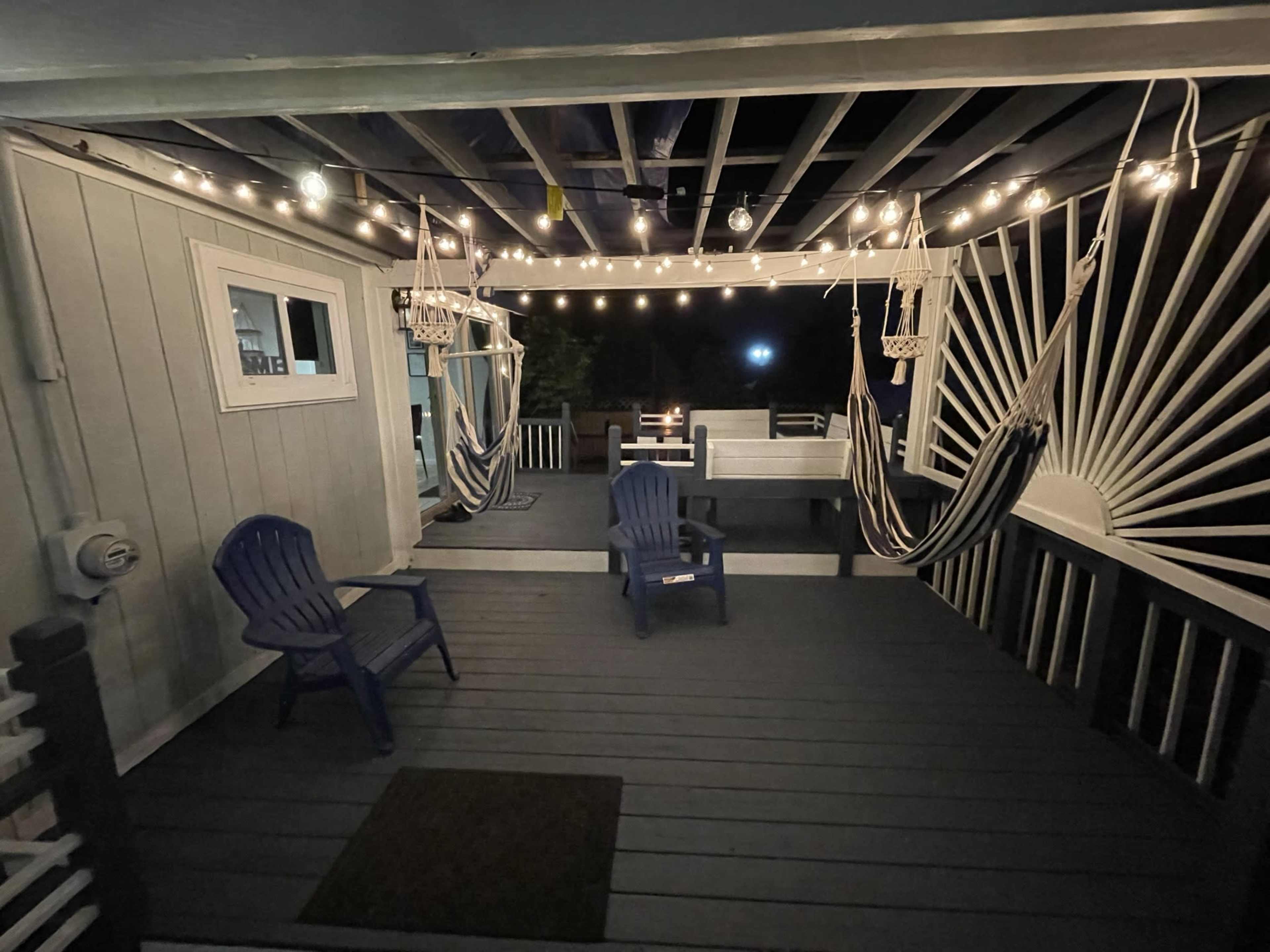 A nighttime view of a porch with hanging lights, two hammocks, and chairs arranged on a wooden floor.