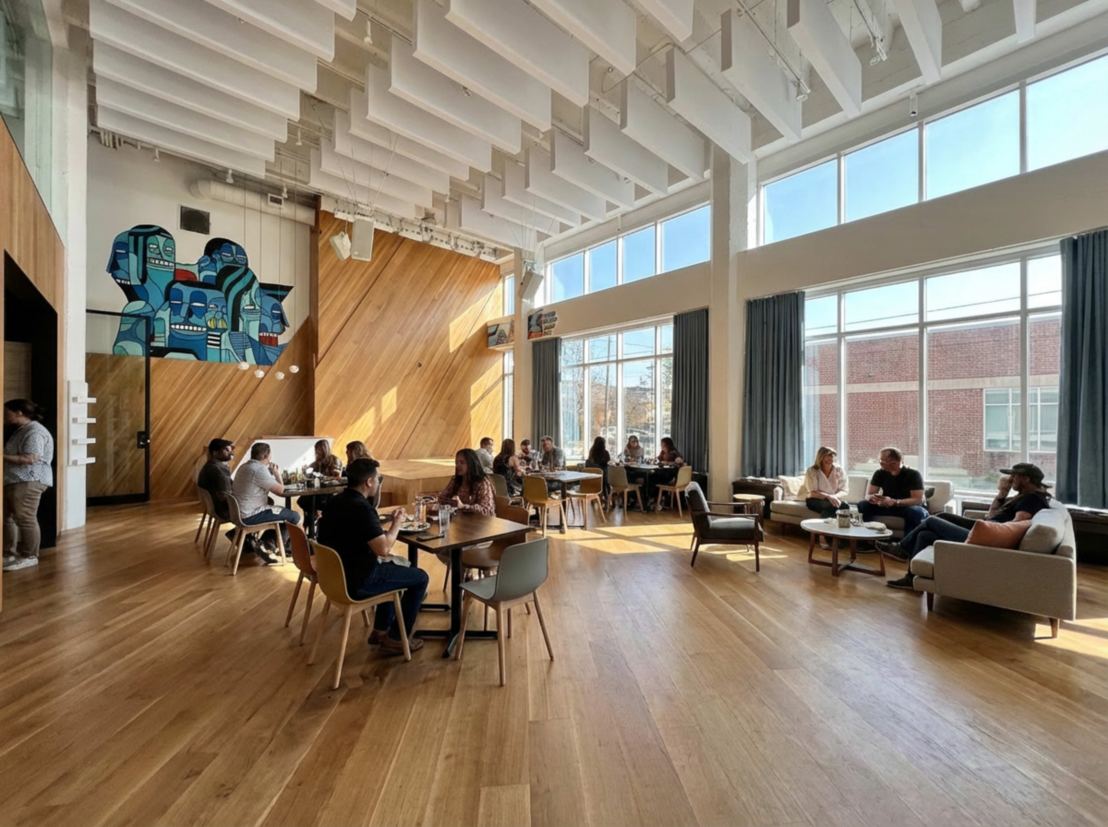 The image shows a modern, well-lit café with large windows, wooden floors, and a mix of seating arrangements, where several groups of people are engaged in conversation or dining.