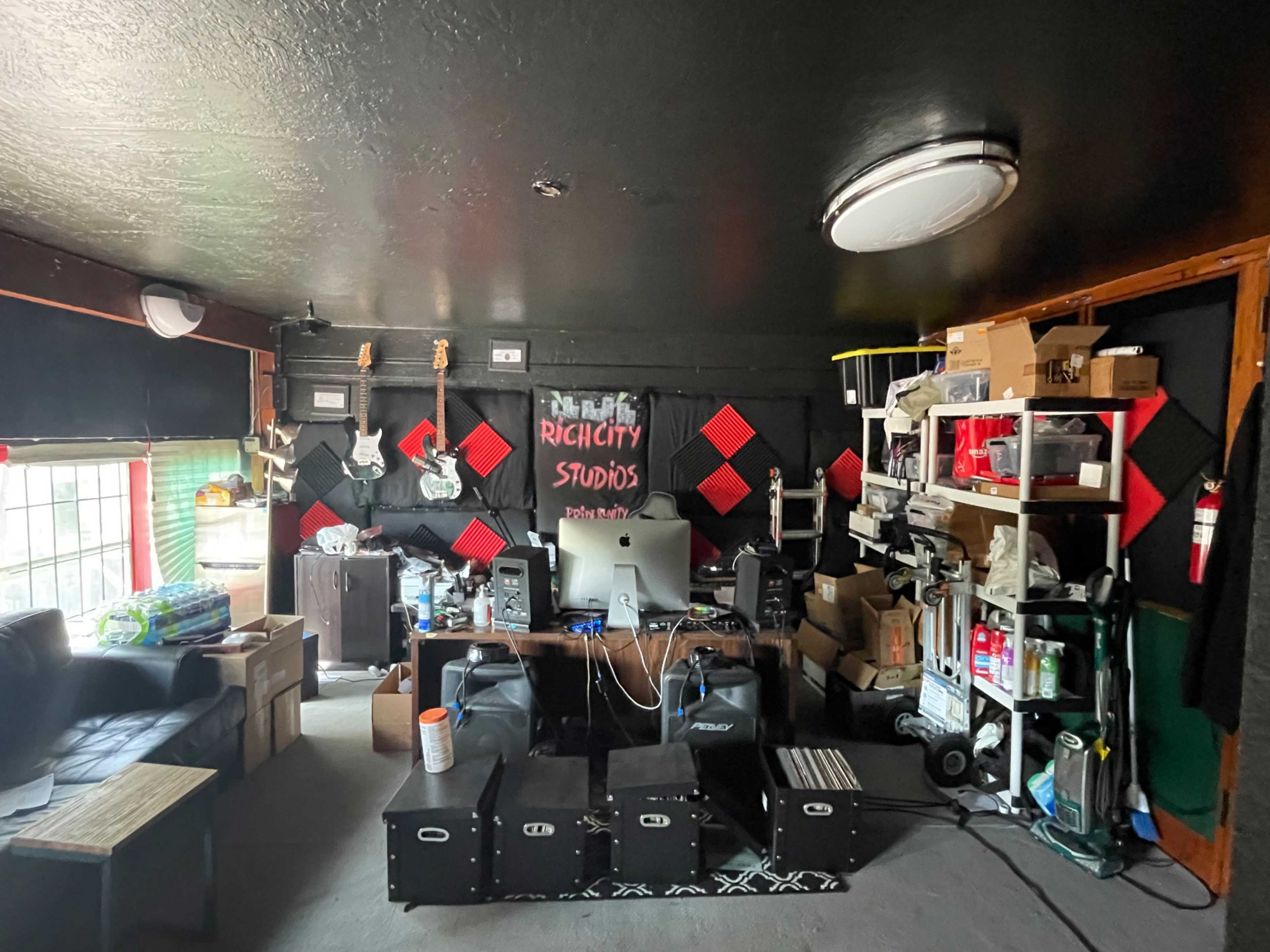 A cluttered music studio featuring instruments on the walls, a computer setup at the center, and shelves filled with various equipment and supplies.