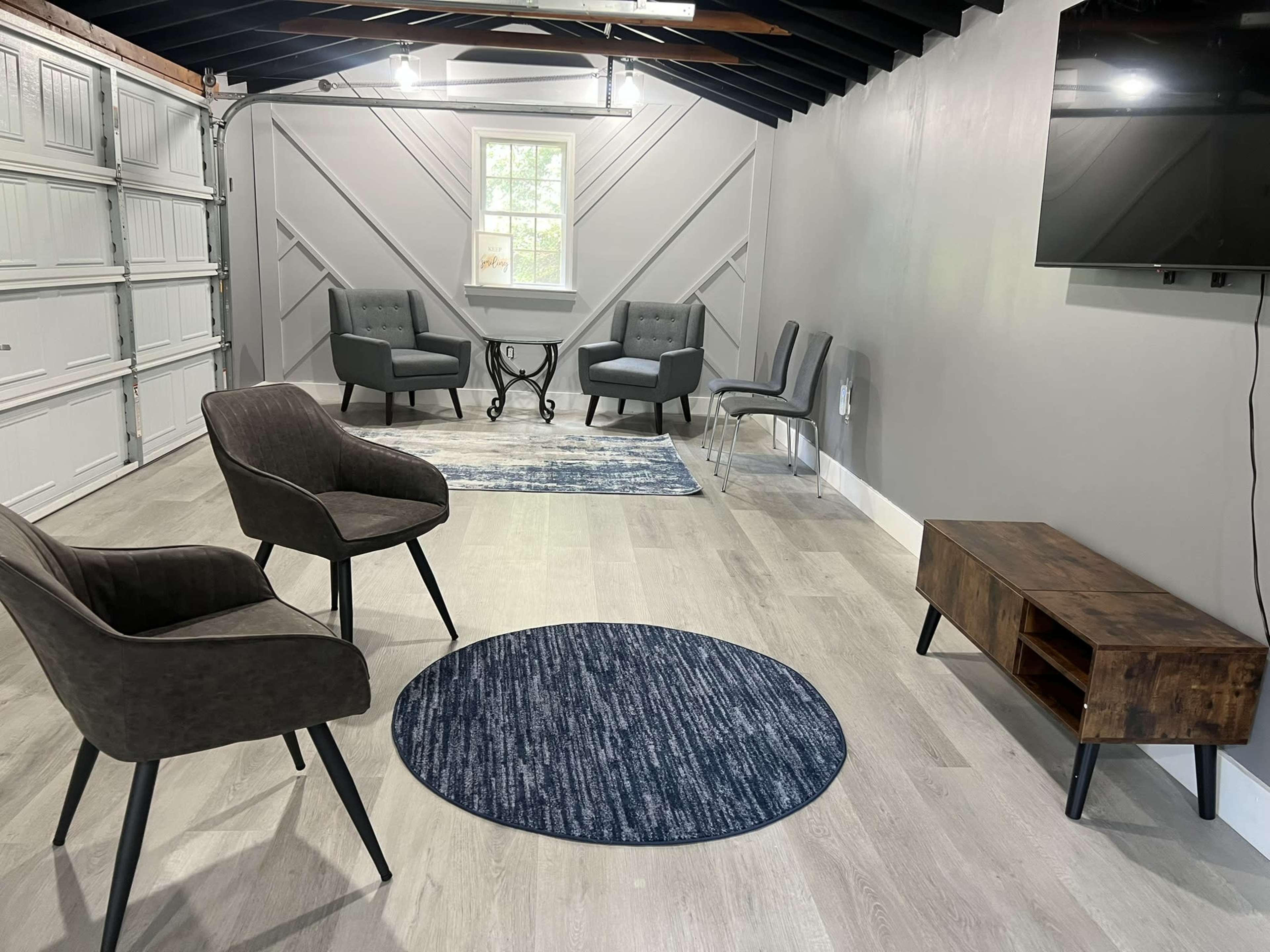 A modern, sparsely furnished room with two types of chairs, a small table, a TV mounted on the wall, and a round rug on the floor.