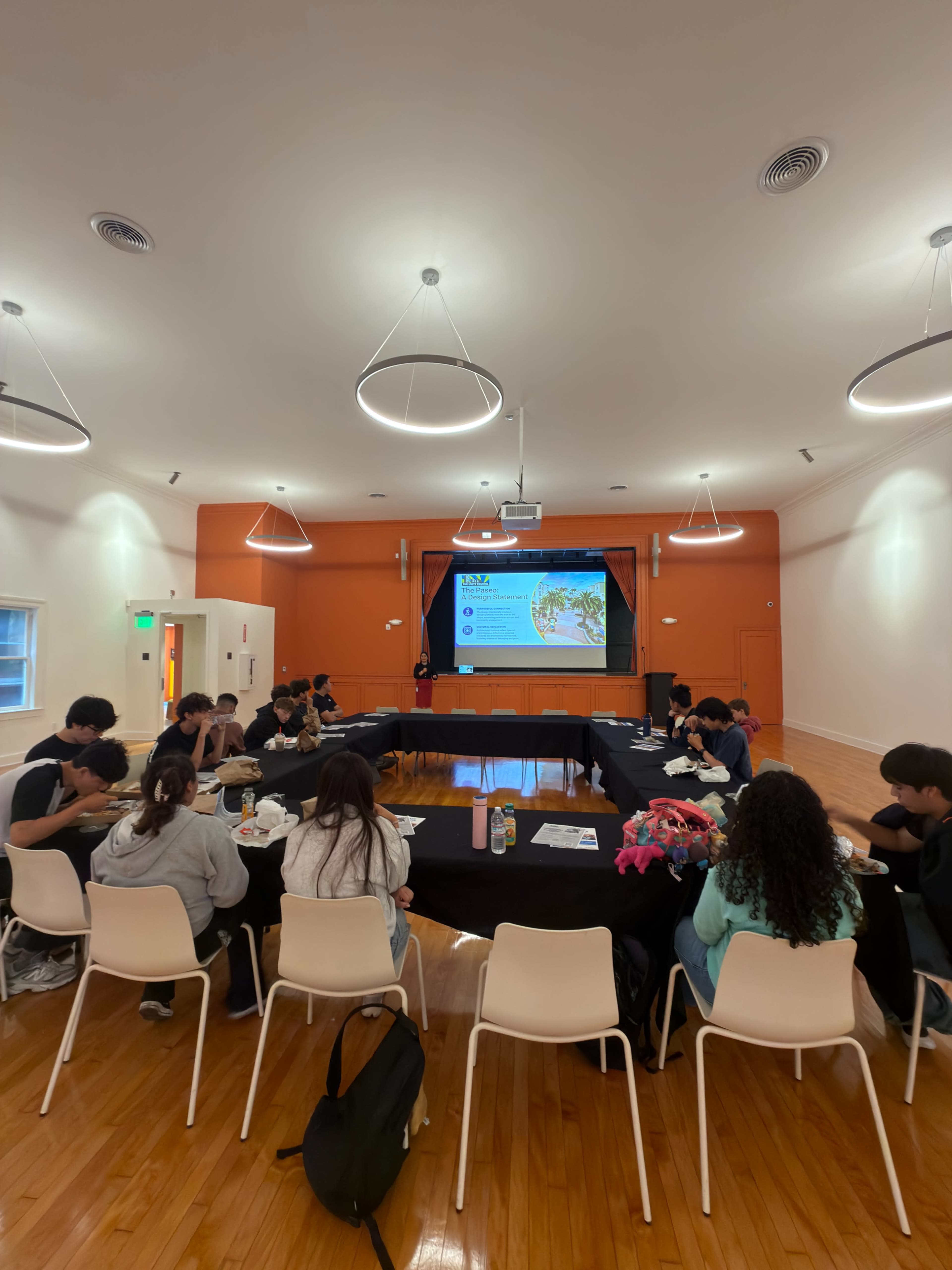 A group of students gathers around a large table in a brightly lit room with an orange accent wall, watching a presentation on a screen.