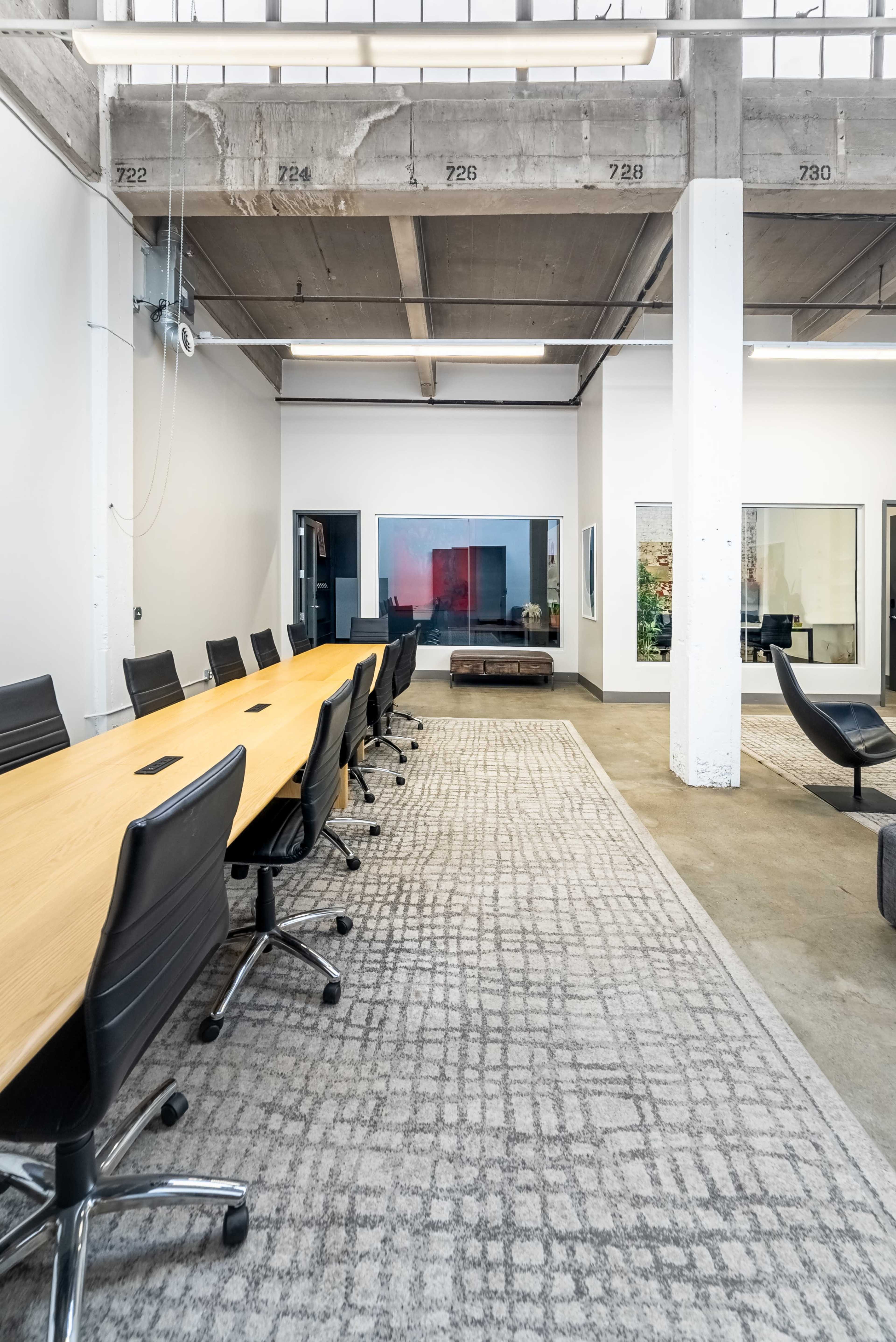 A long wooden conference table with rolling chairs is set in a modern office space featuring large windows and a patterned rug.