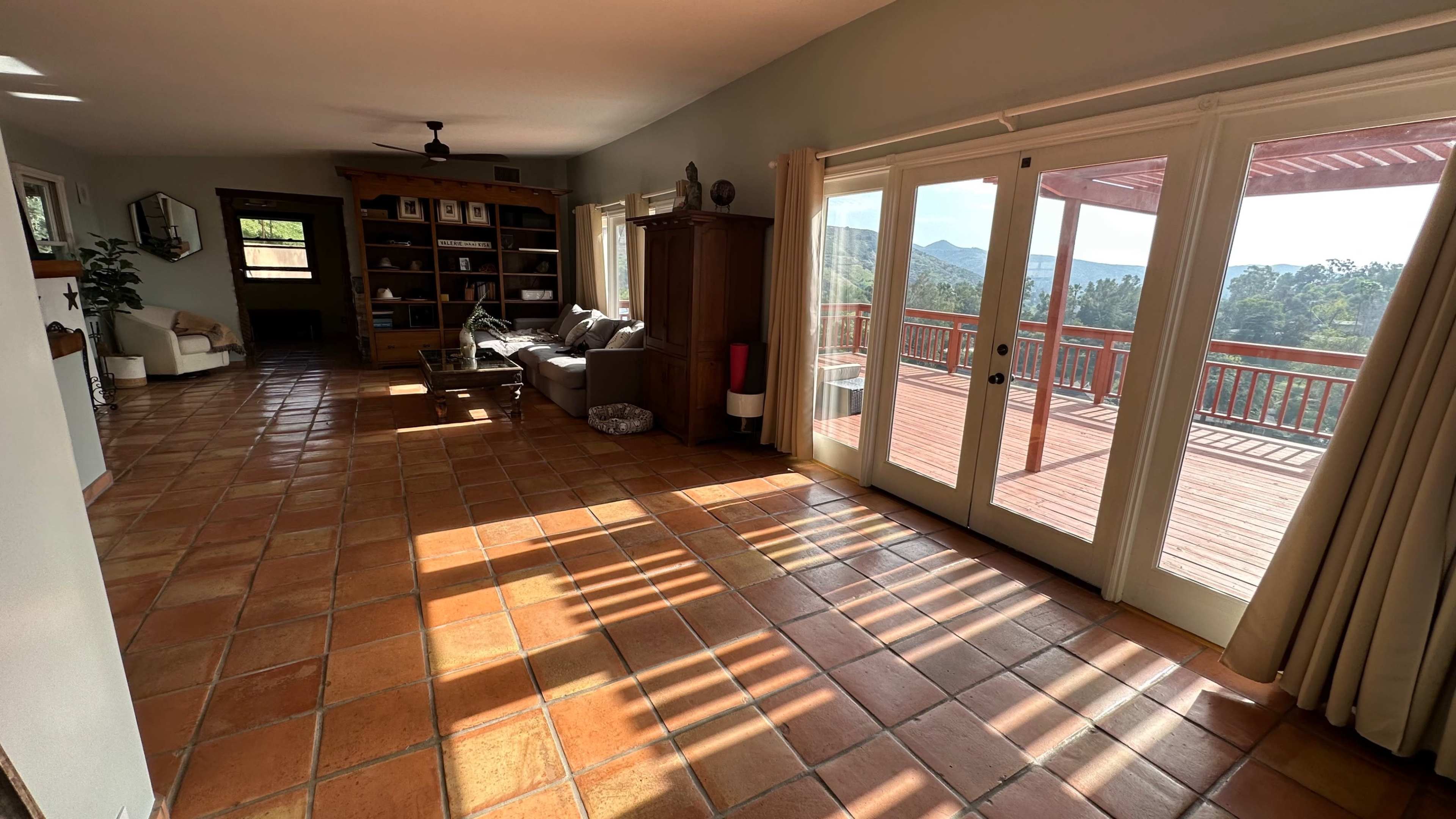 The spacious living room features a tiled floor, large windows with sunlight casting shadows, and opens onto a deck with mountain views.