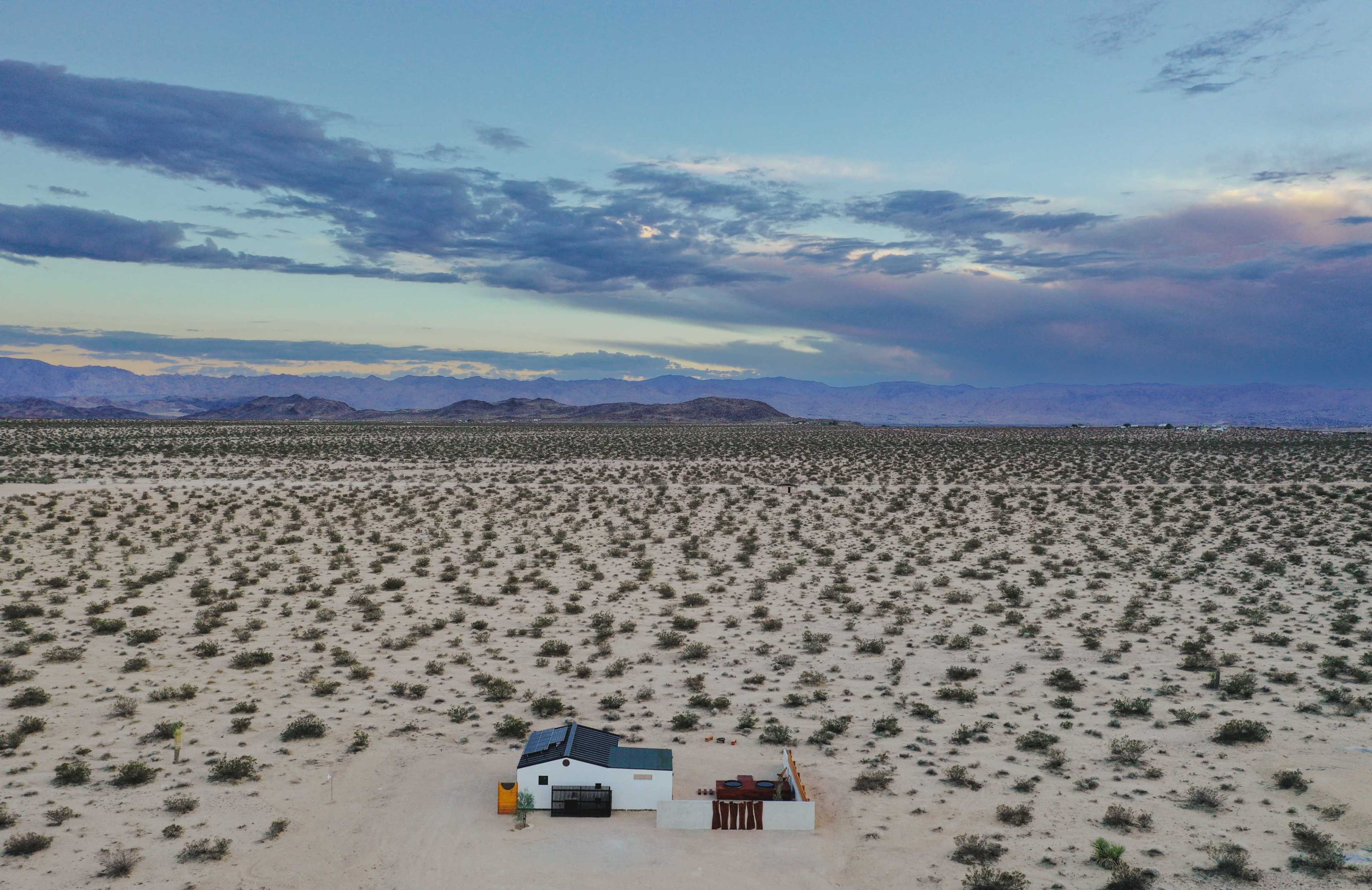 A small white house sits on an expanse of dry desert land surrounded by low vegetation and distant mountains under a cloudy sky.