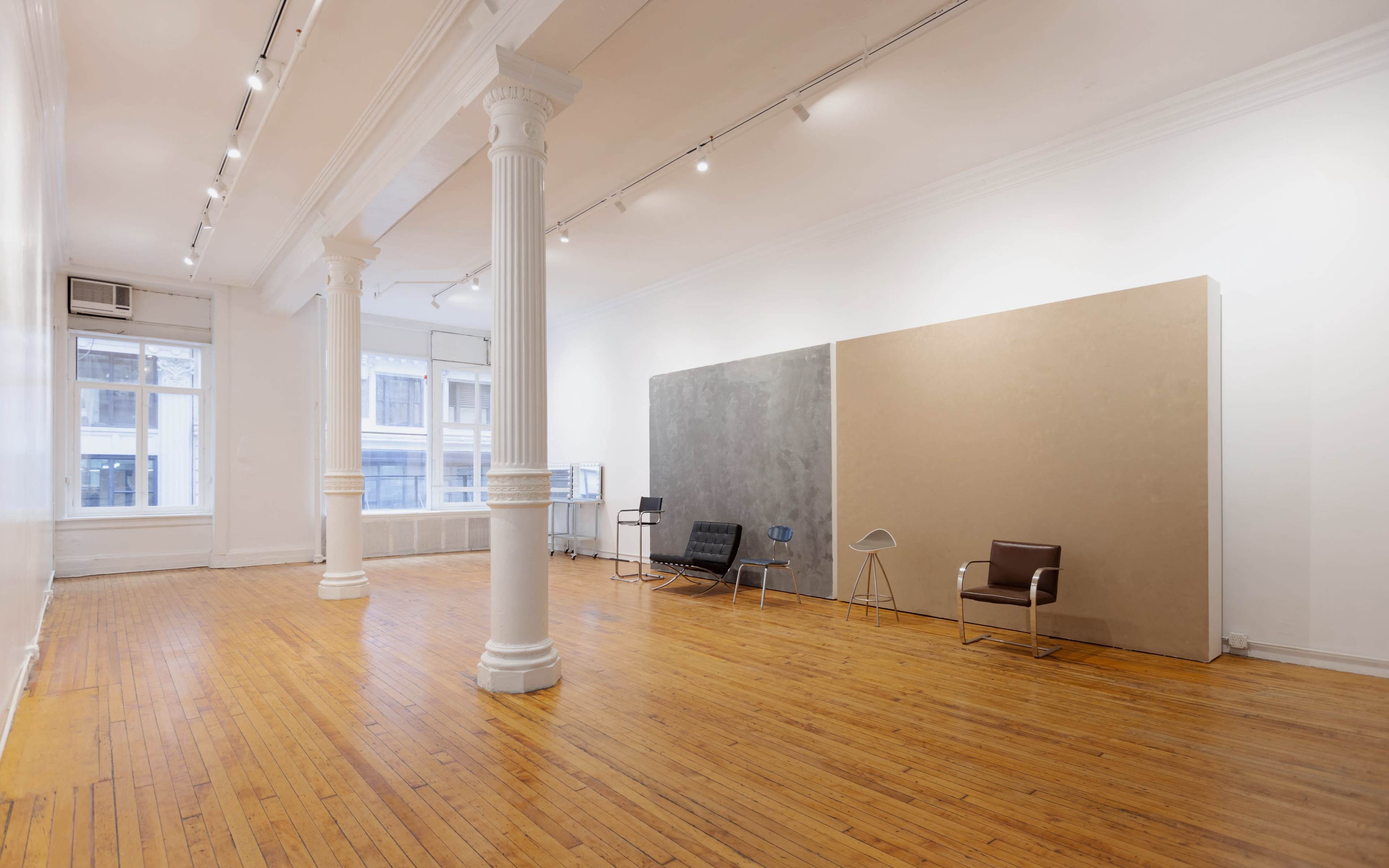 The image shows a spacious, empty art gallery with wooden floors, two large pillars, and minimalist furniture against blank walls.