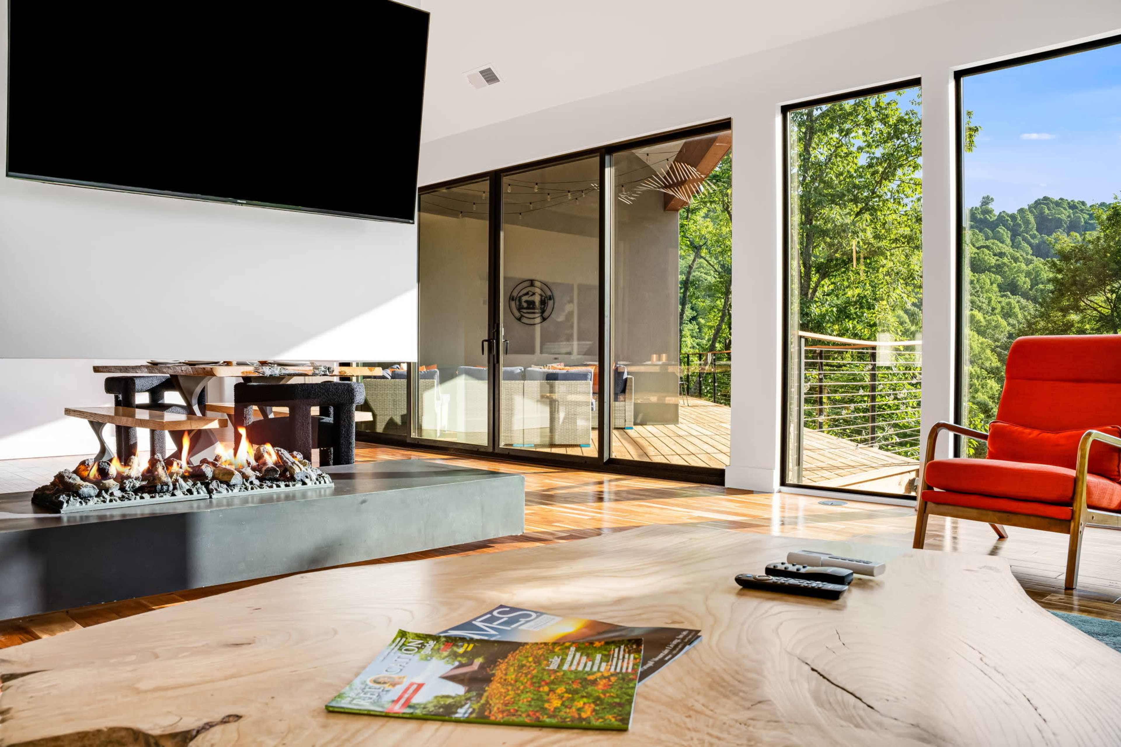 A modern living room features a fireplace, a large TV mounted on the wall, floor-to-ceiling windows offering a view of the outdoors, and a red armchair next to a wooden coffee table with magazines.