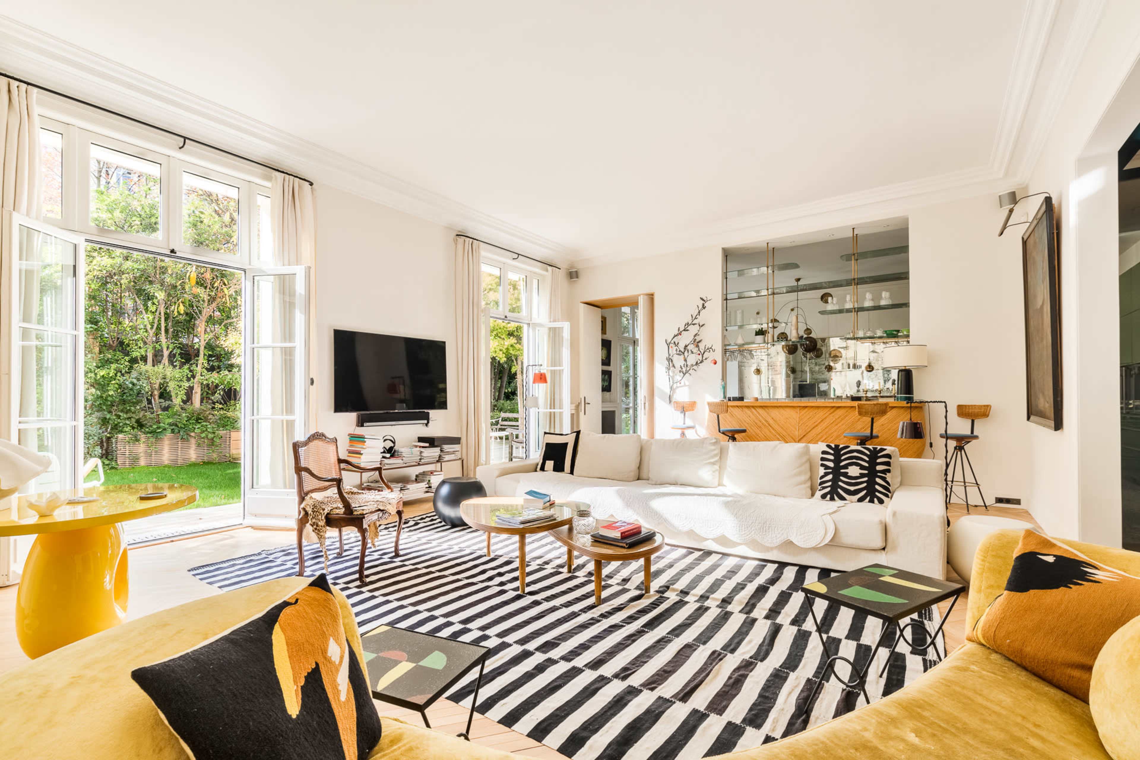 The image shows a bright living room with large windows, a comfortable sofa, a striped rug, and a wooden sideboard displaying decorative items.