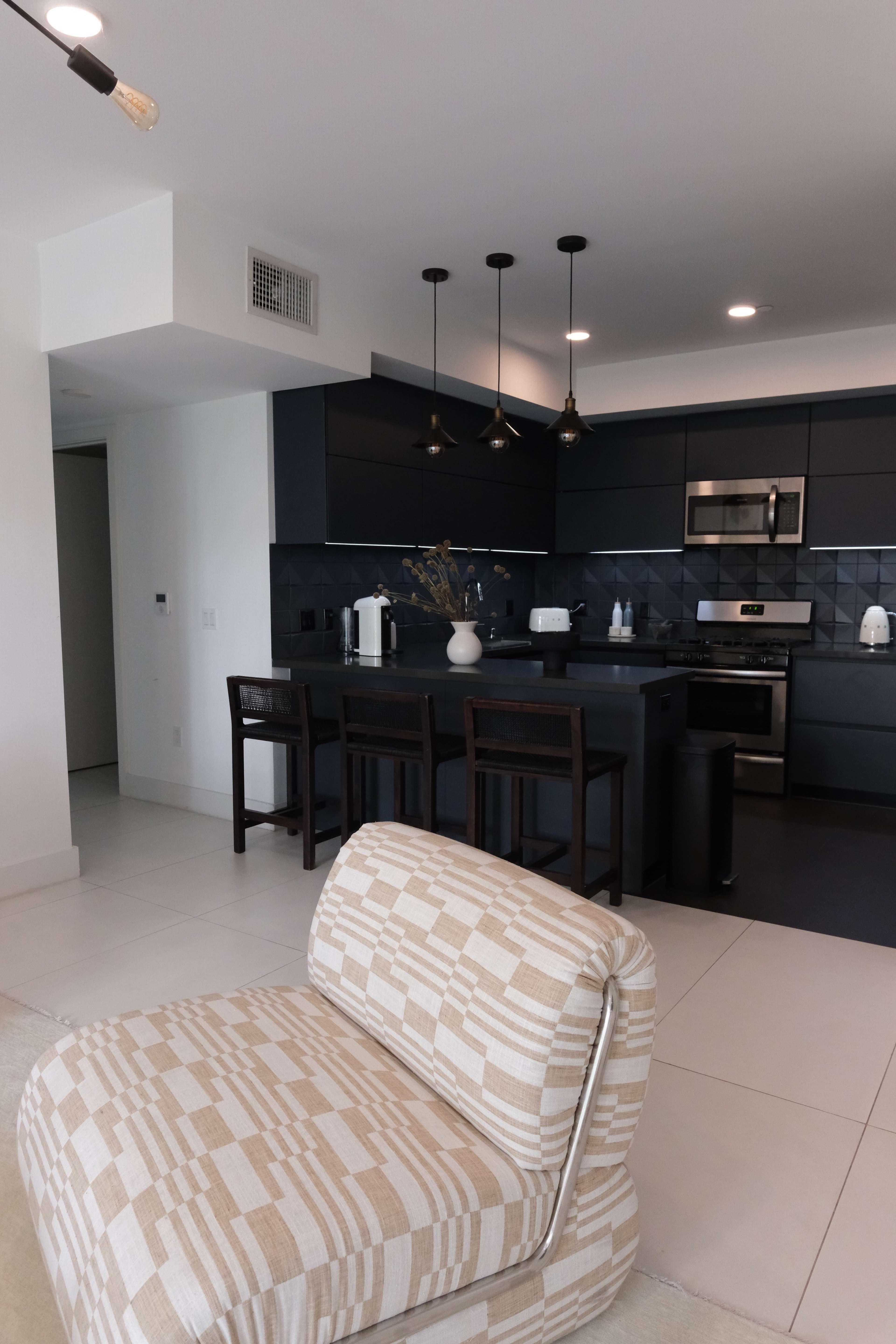 The image shows a modern kitchen with dark cabinetry, stainless steel appliances, and three pendant lights above a bar counter with four stools.