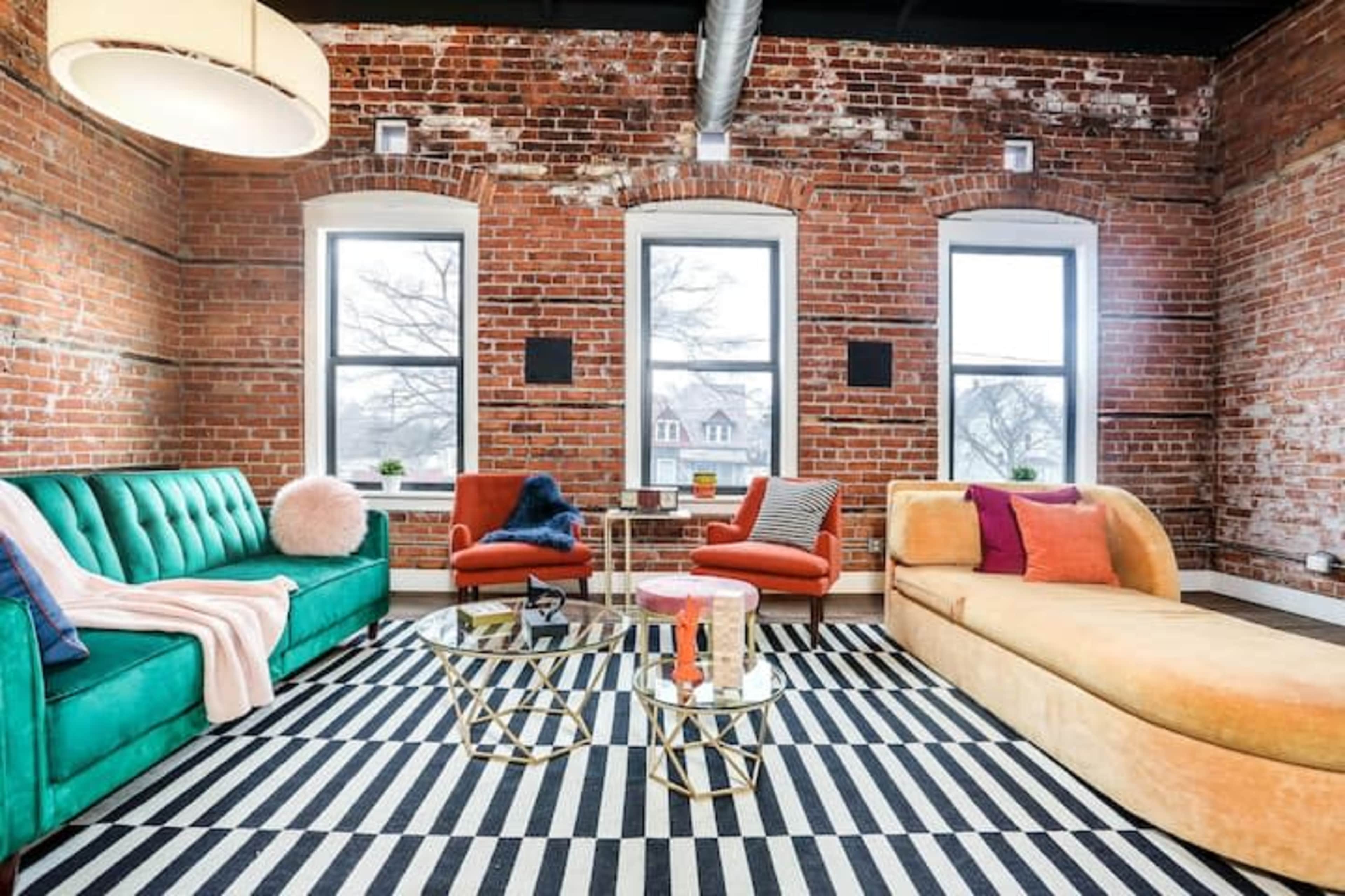 A stylish living room features a green sofa, orange chairs, and a gold and glass coffee table set against a backdrop of exposed brick walls and large windows.