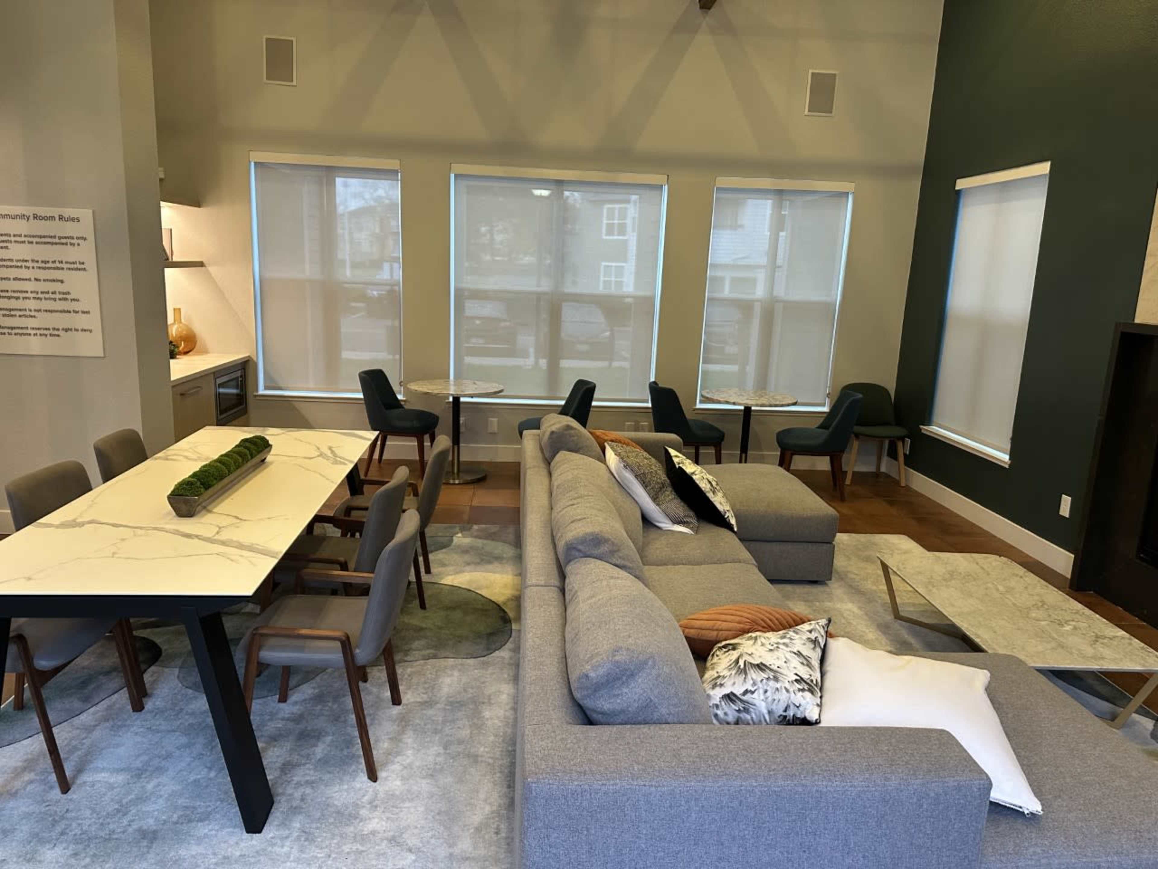 A modern communal lounge featuring a large gray sectional sofa, a marble dining table with chairs, and large windows allowing natural light.