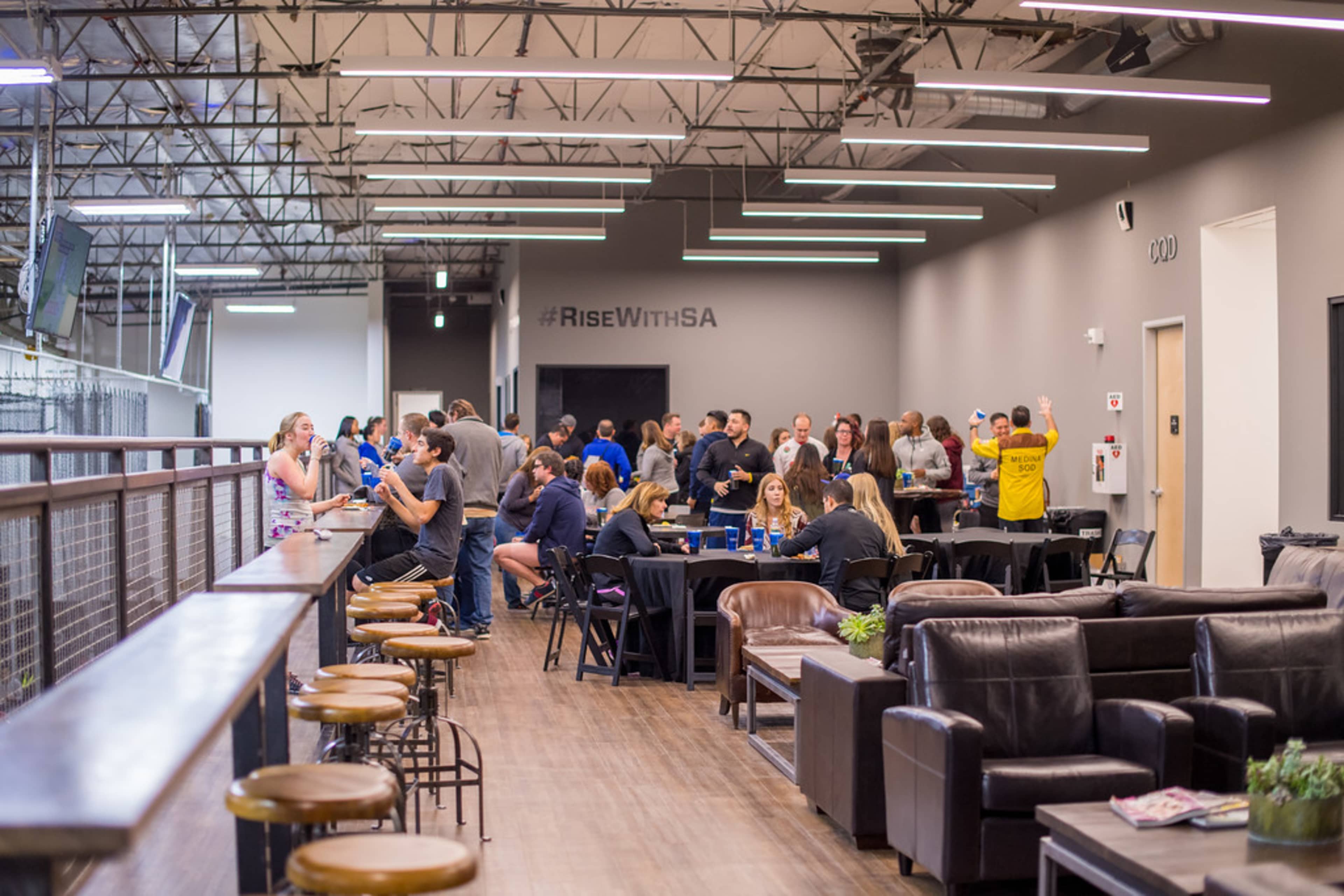 A group of people socializes in a spacious indoor venue with tables and seating, while a large wall displays the hashtag "#RiseWithSA."