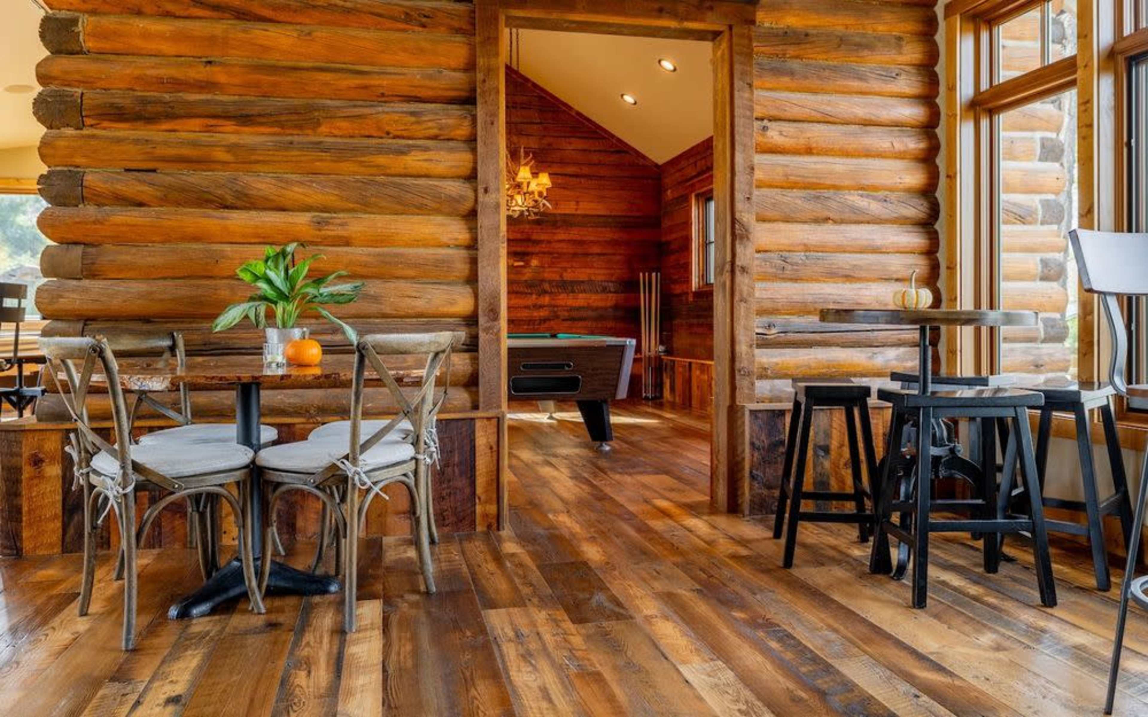 The image shows a rustic interior space with log walls, a wooden floor, and a view into a room with a pool table.