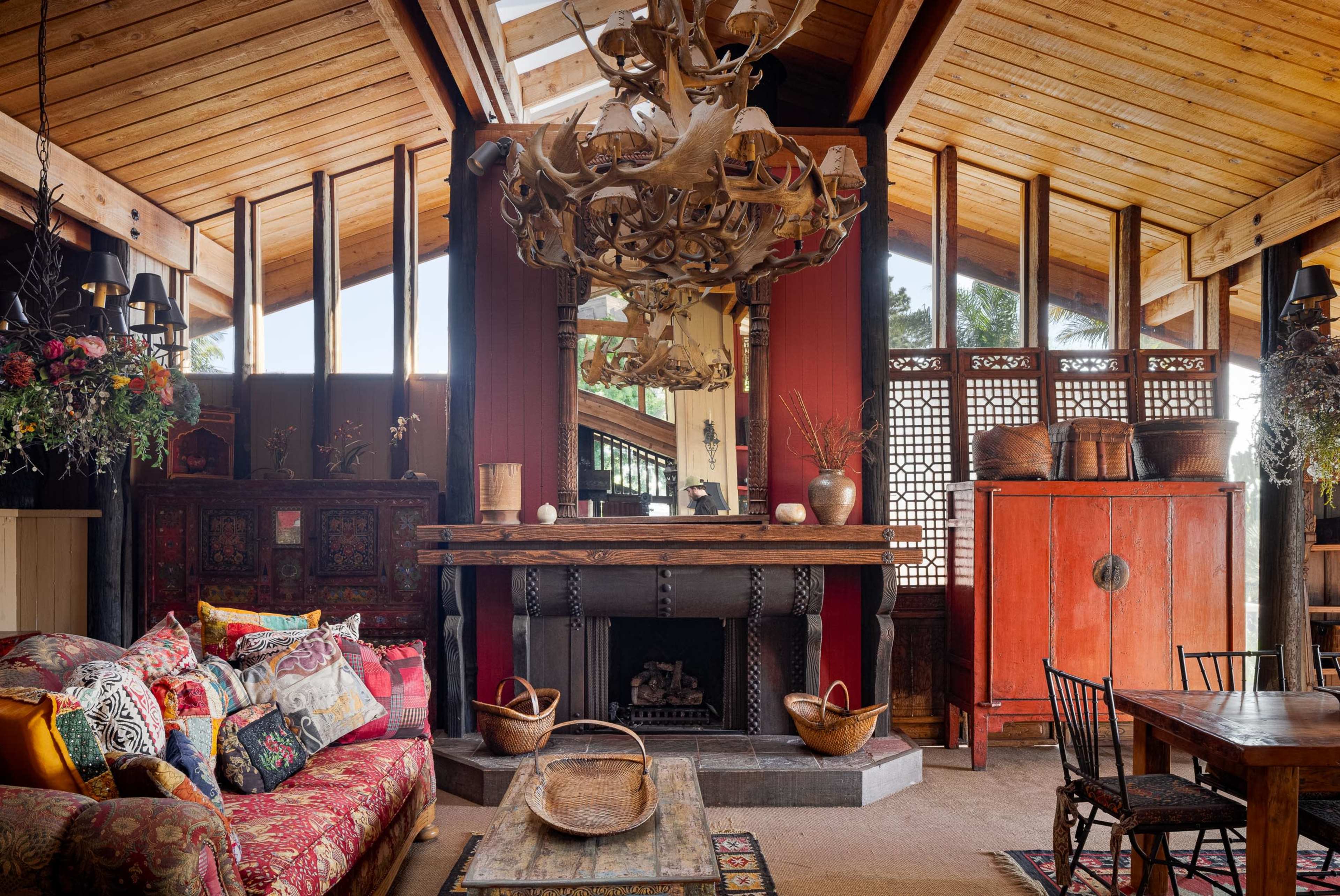 The image shows a cozy living space featuring a decorative sofa, a large wooden fireplace, and rustic furnishings under a wooden ceiling with a chandelier.