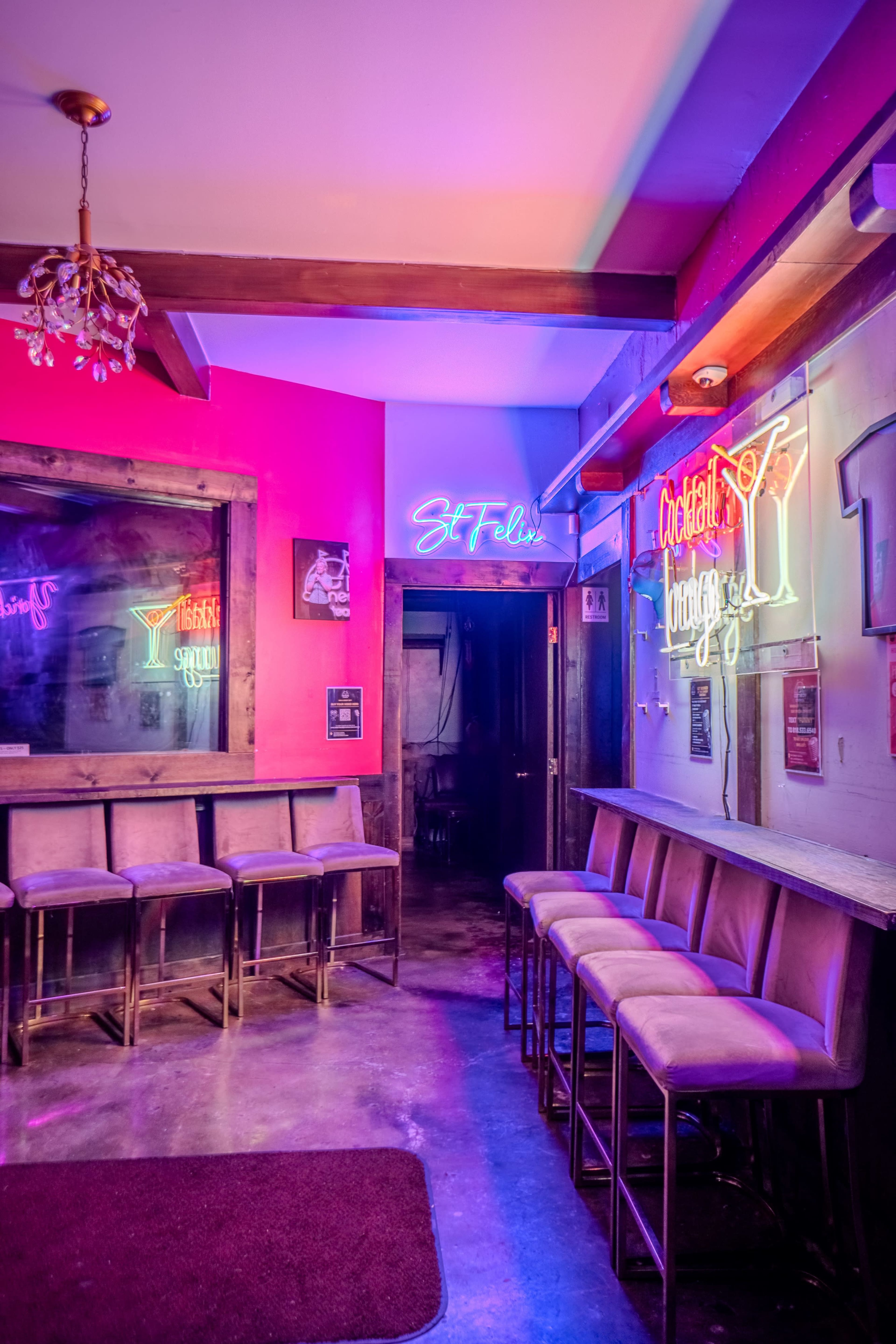 The interior of a bar features a row of stools against a wall, illuminated by colorful neon signs and soft lighting.