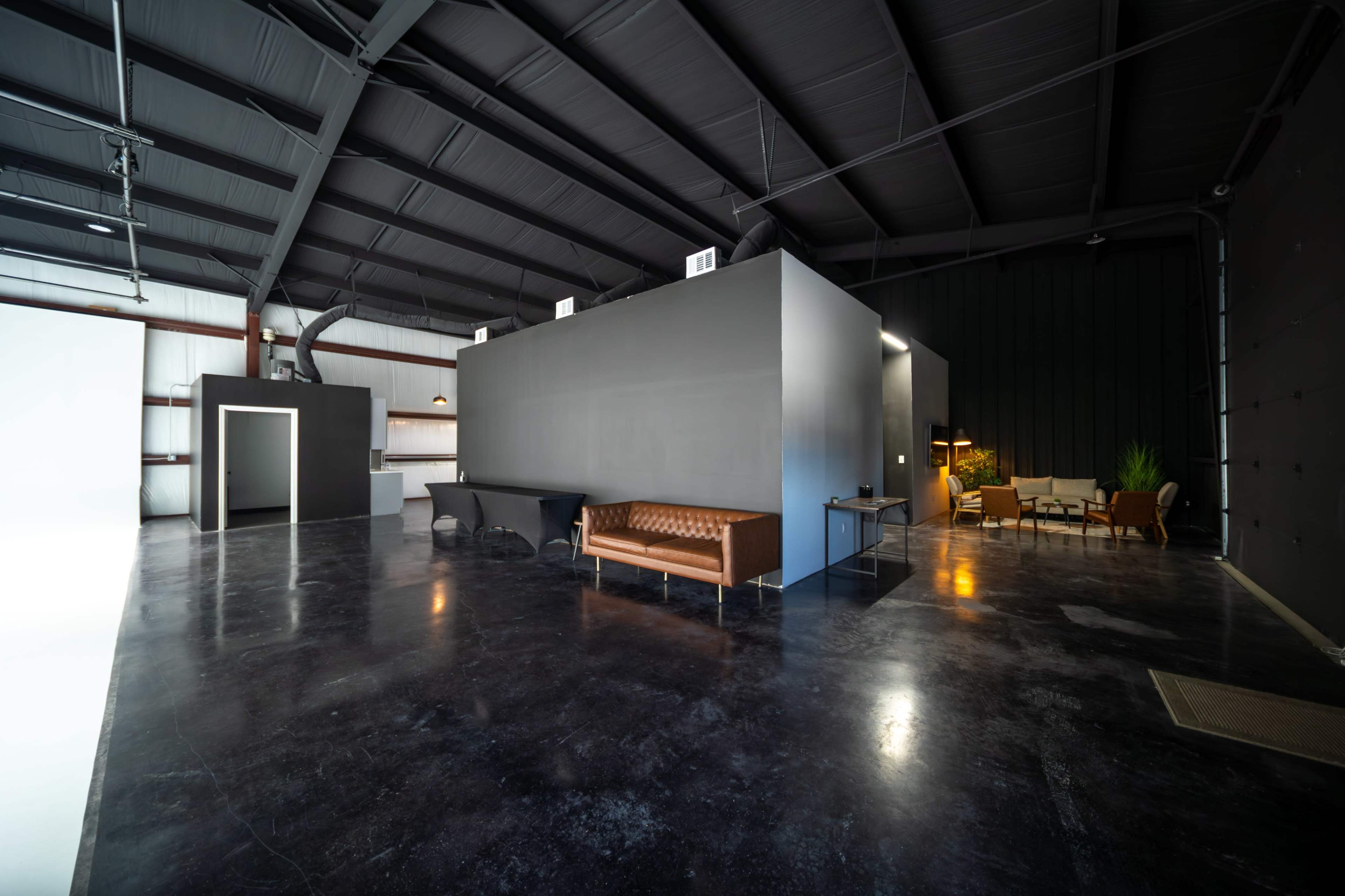 The image shows a spacious industrial studio with polished concrete floors, featuring a central gray structure, a leather sofa, and various seating areas along the walls.