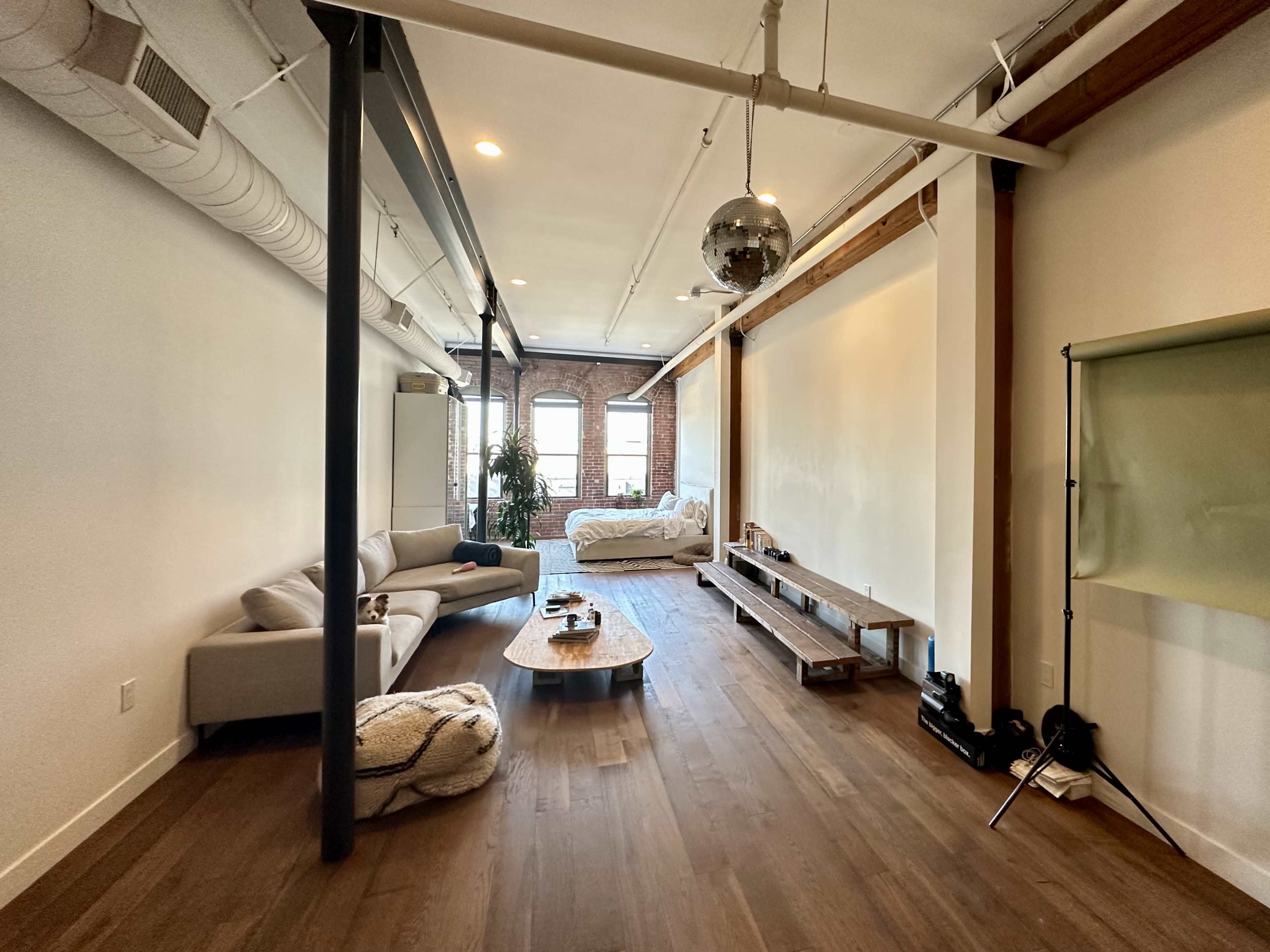 A spacious, minimalistic living room features a light-colored sofa, wooden floor, and a large window with a view.