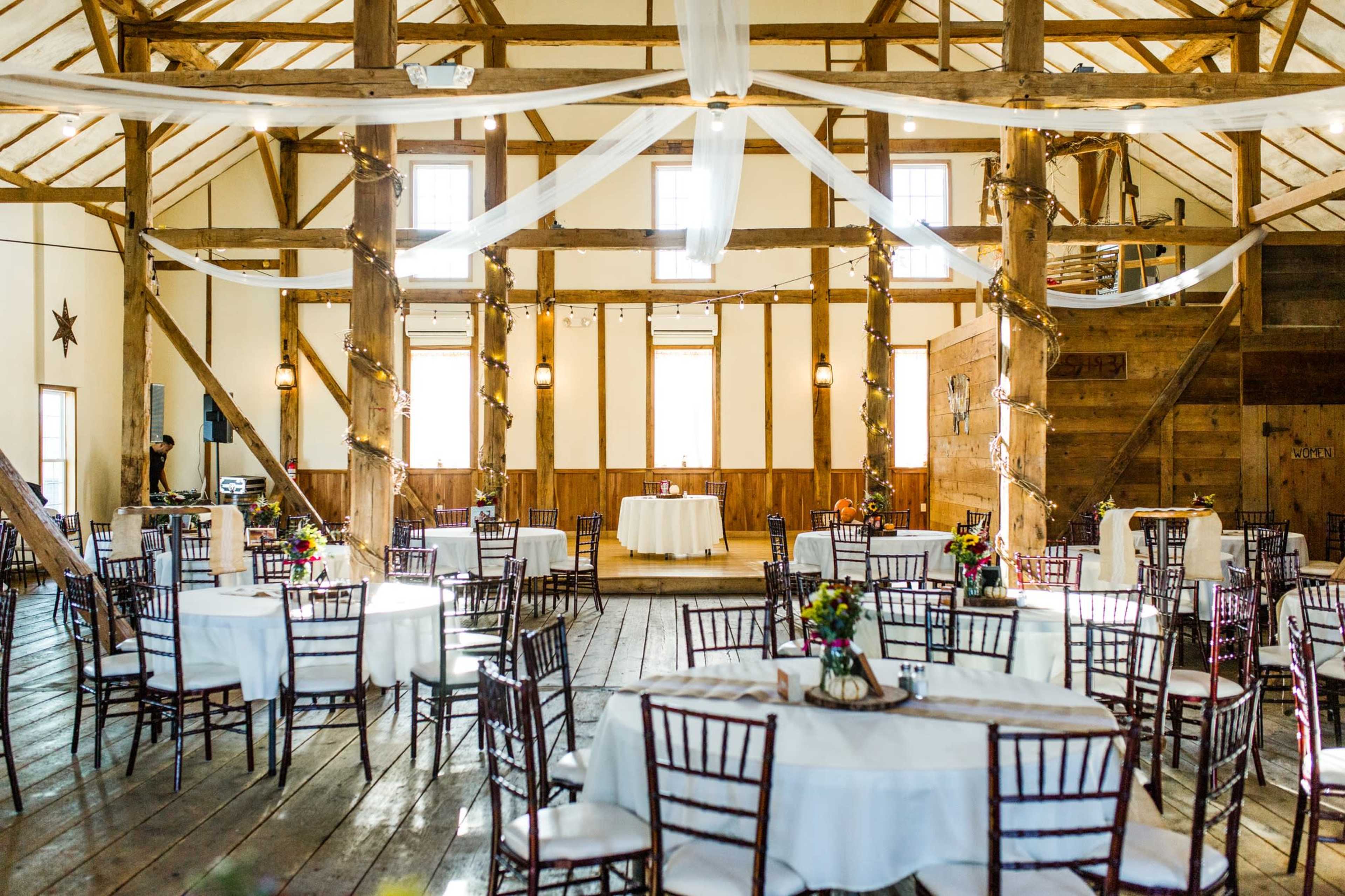 A spacious barn interior features round tables with white tablecloths and floral centerpieces, surrounded by wooden beams and draped fabric.