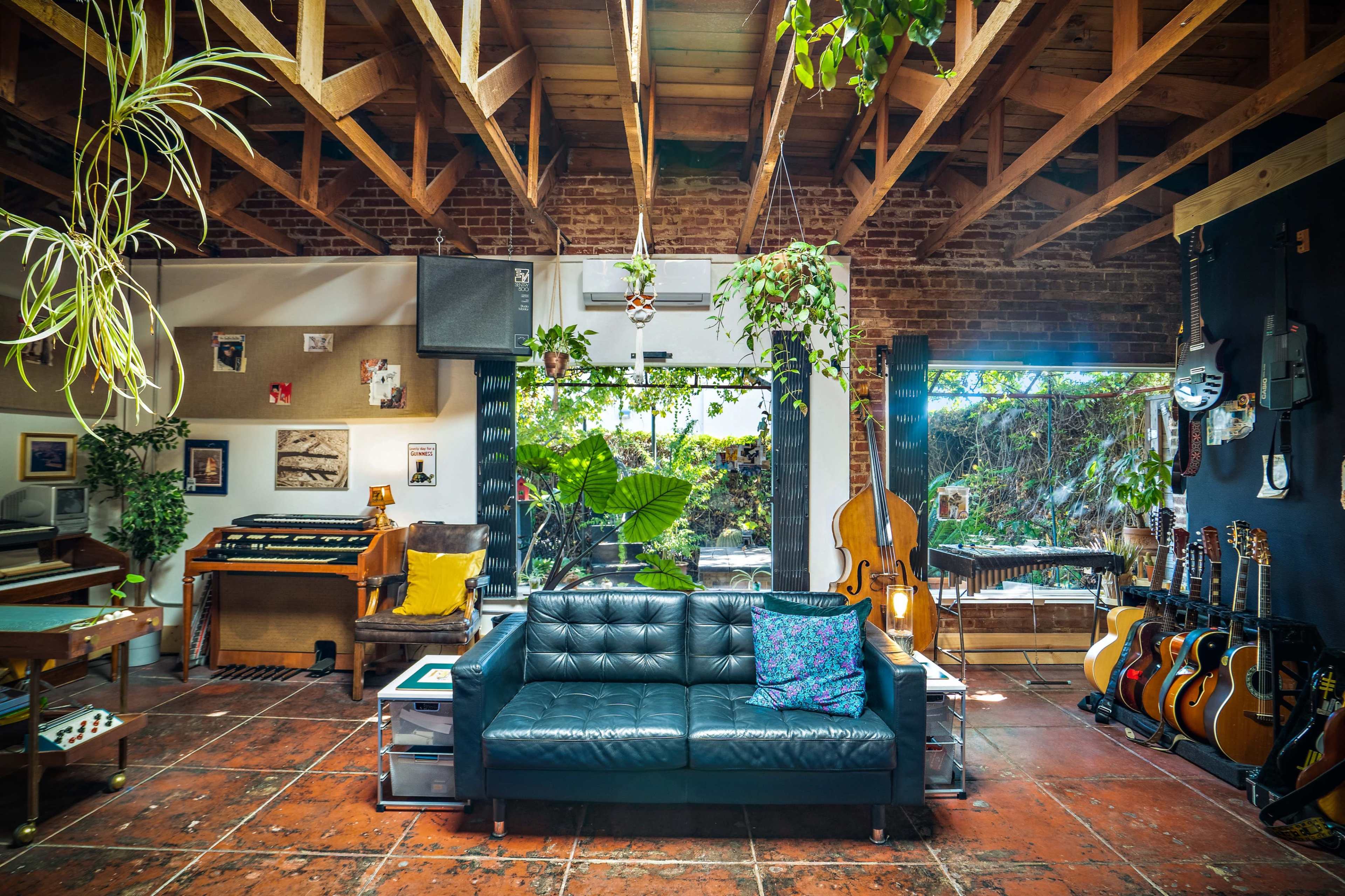 A cozy living room features a black leather couch, musical instruments, and large windows overlooking a green garden.