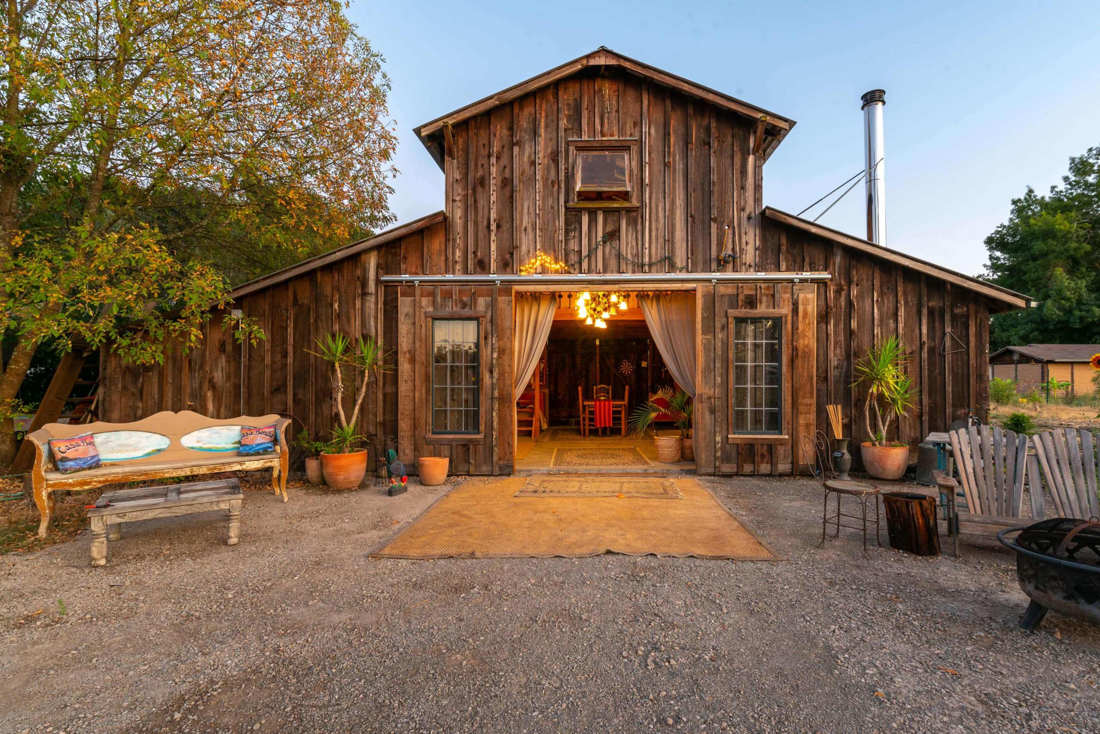 A rustic wooden barn with large open doors reveals a warmly lit interior, surrounded by plants and outdoor seating.