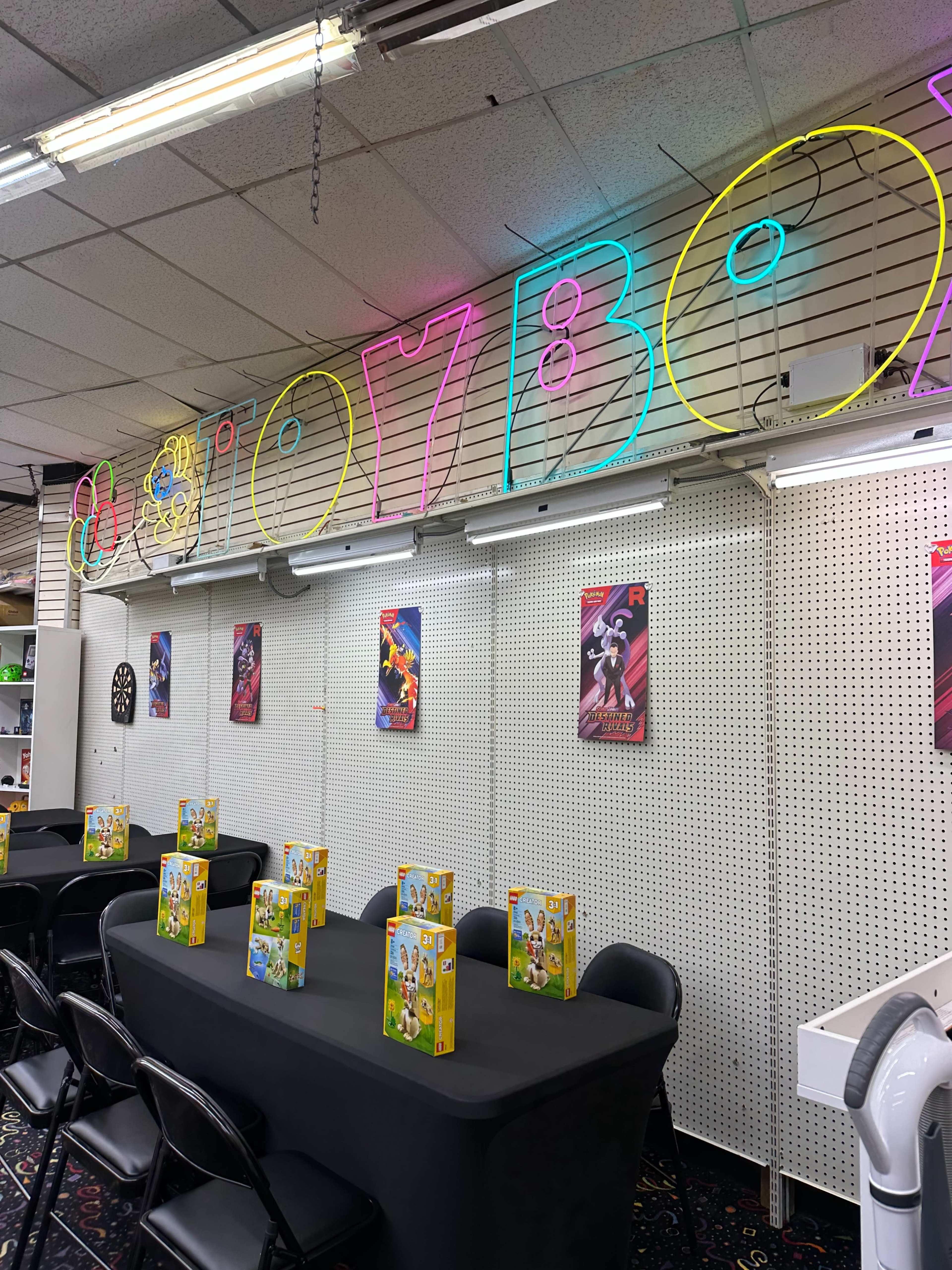 The image shows a brightly lit gaming room with a neon "GAMING BOX" sign, black tables set for games, and colorful posters on the walls.