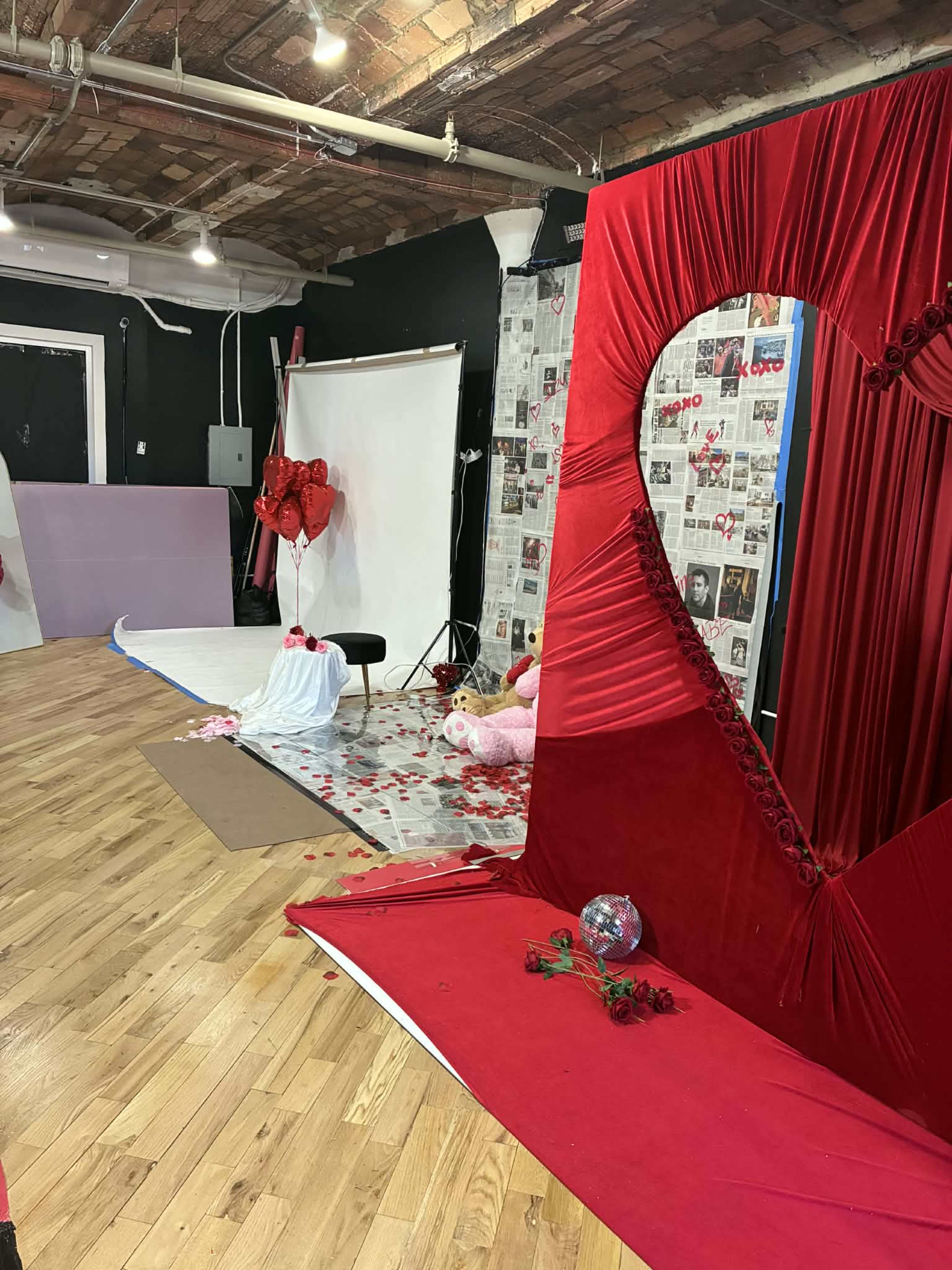 The image shows a photography studio set up with a red heart-shaped backdrop, pink and white decorations, and a variety of props scattered on the wooden floor.