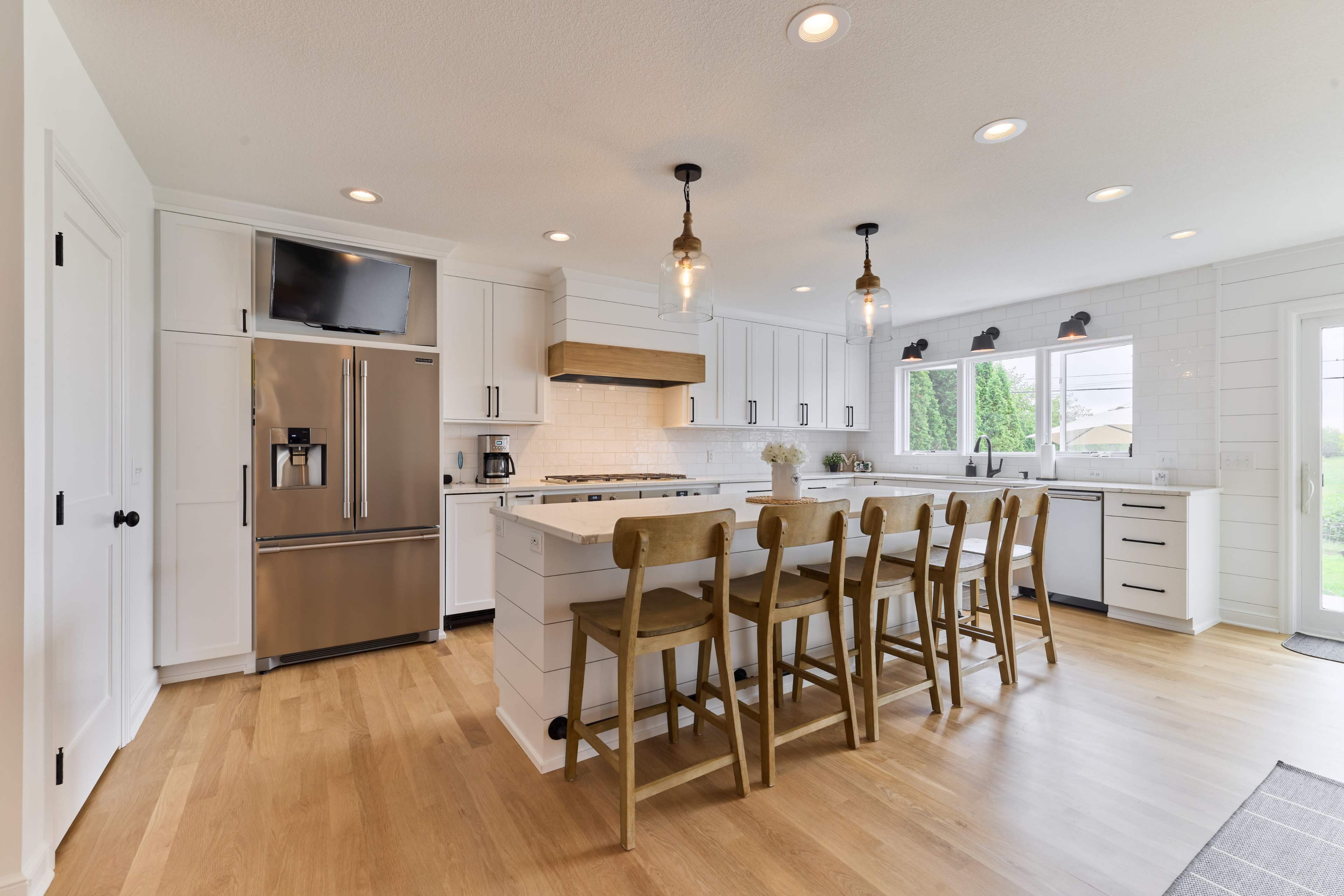 A modern kitchen features white cabinetry, a large island with seating, stainless steel appliances, and windows overlooking a yard.
