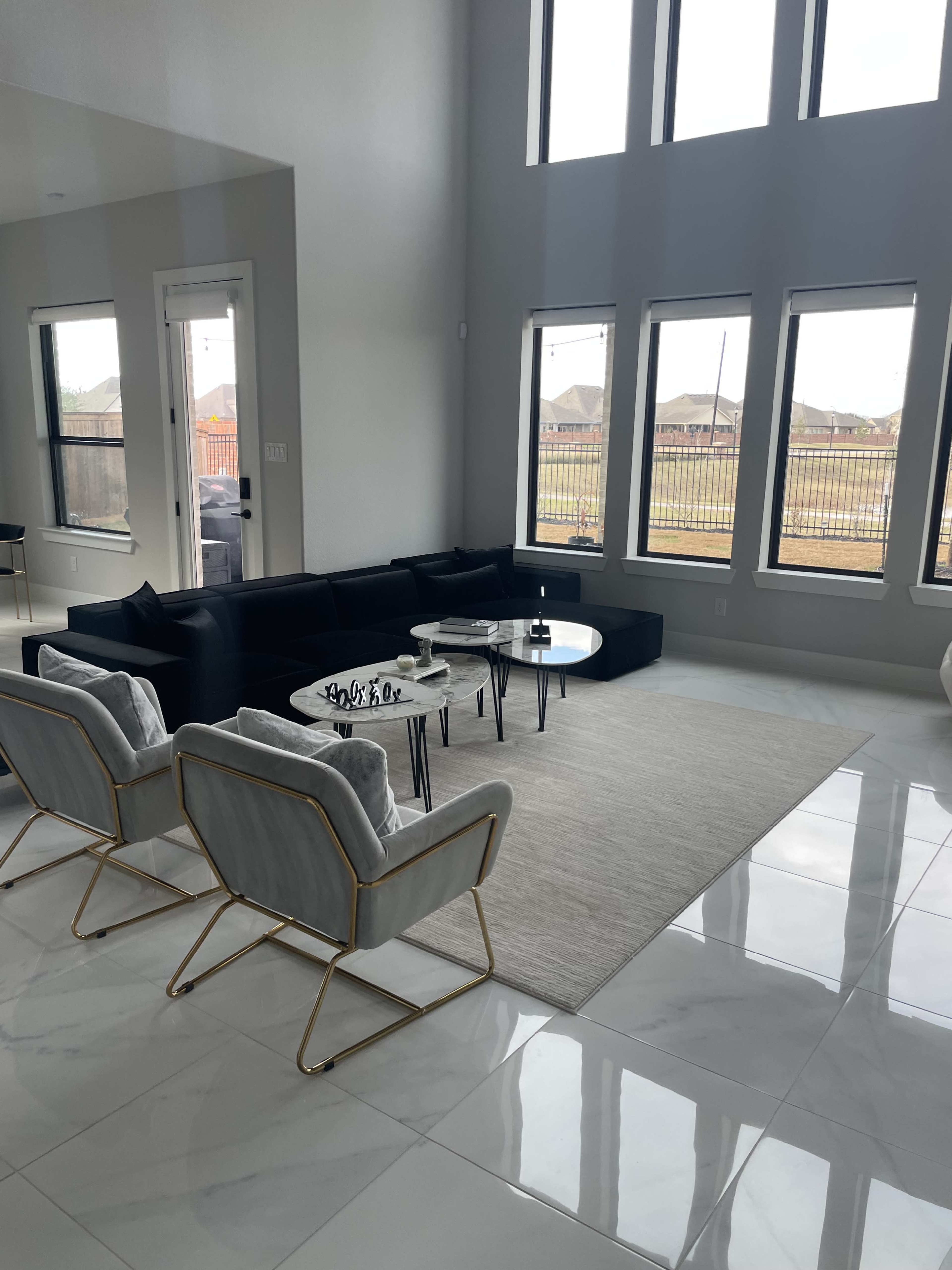 A modern living room with large windows, a black sectional sofa, two light gray chairs, and a round coffee table on a light-colored rug over glossy white tiles.