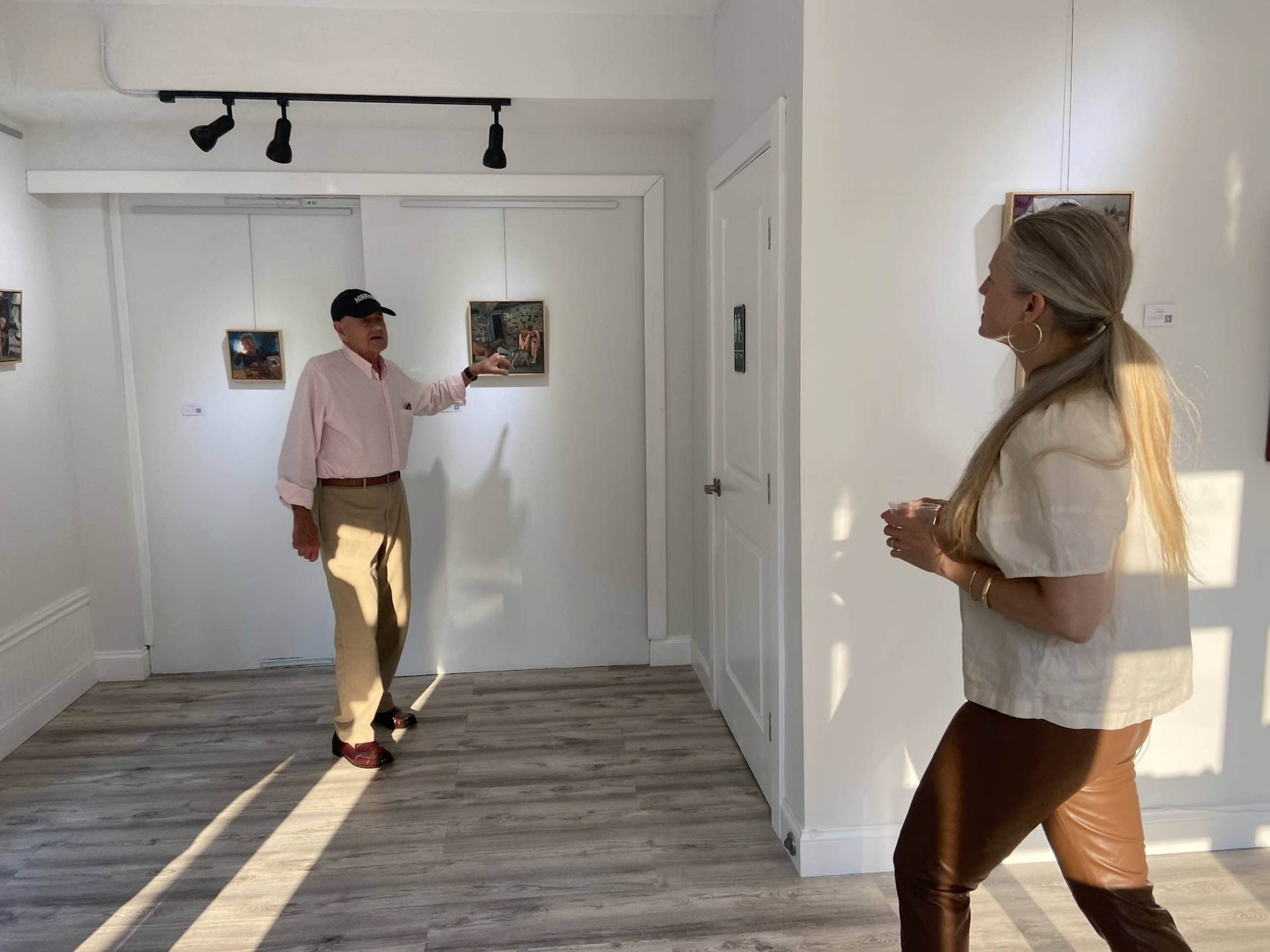 A man in a pink shirt gestures towards artwork on a gallery wall while a woman in a white top and brown pants listens.