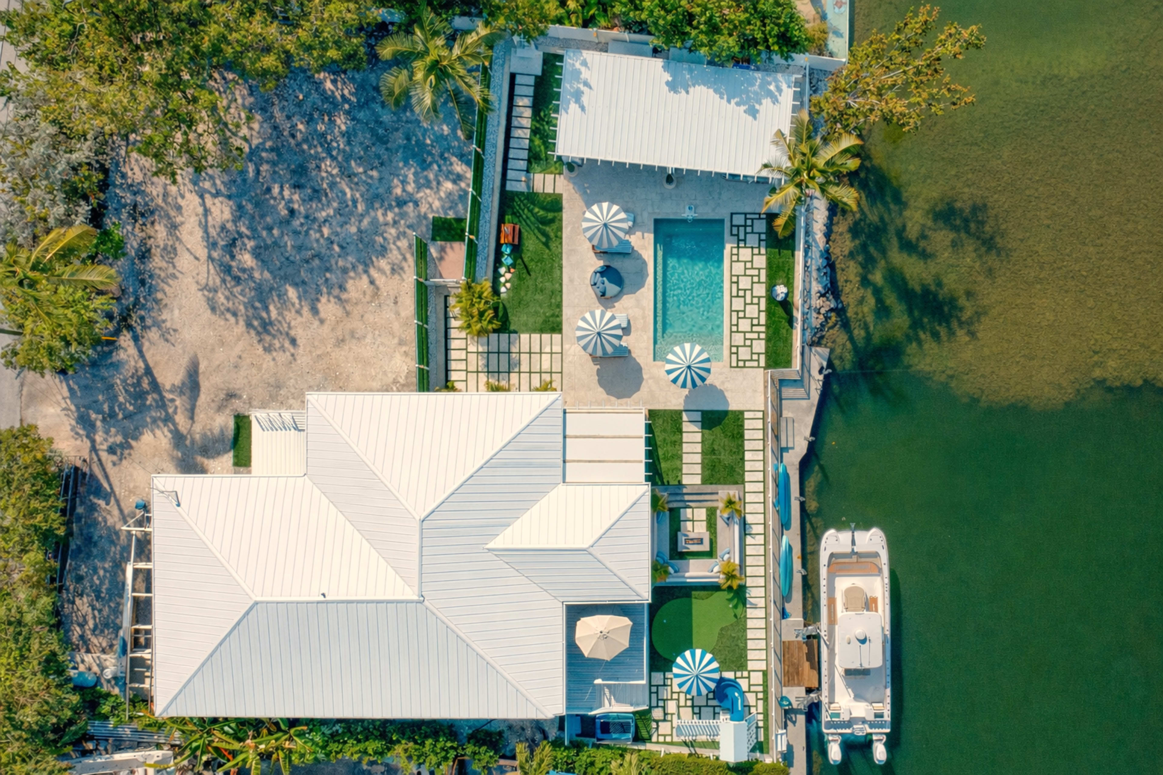 An aerial view captures a modern waterfront house with a pool, outdoor seating areas, and landscaping adjacent to a waterway.