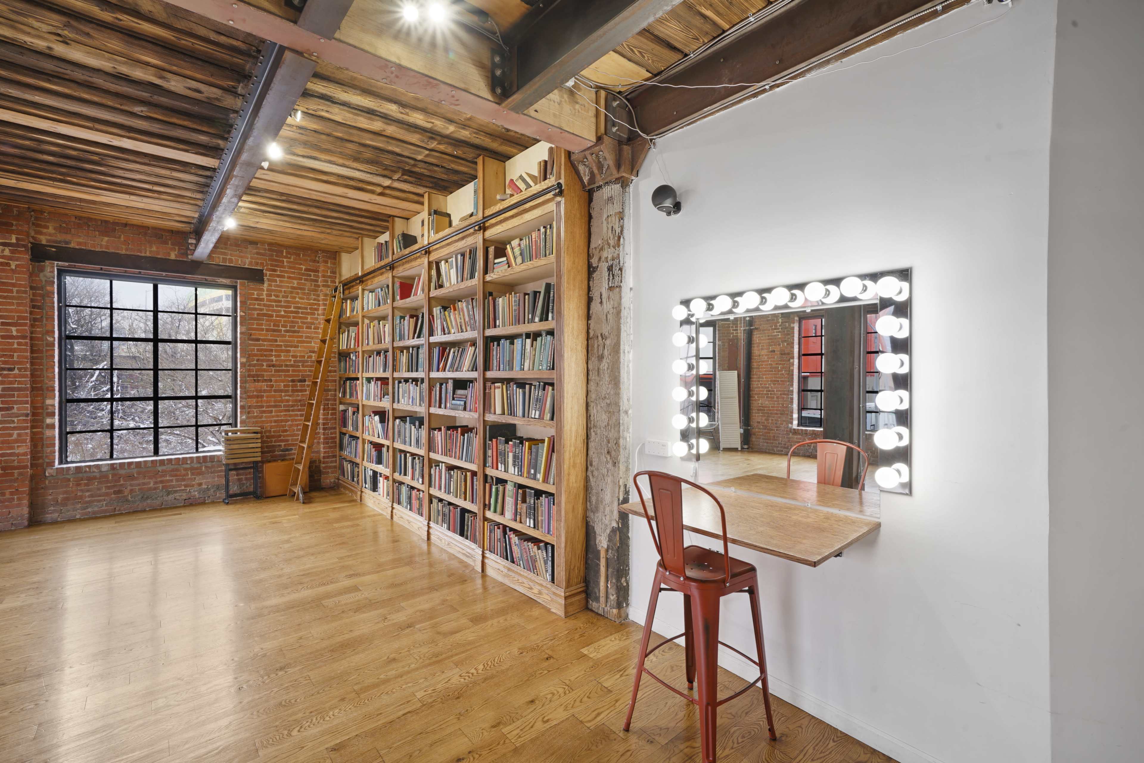 The image shows a spacious room with exposed brick walls, a tall bookshelf filled with books, and a vanity mirror with lights beside a wooden table and chair.