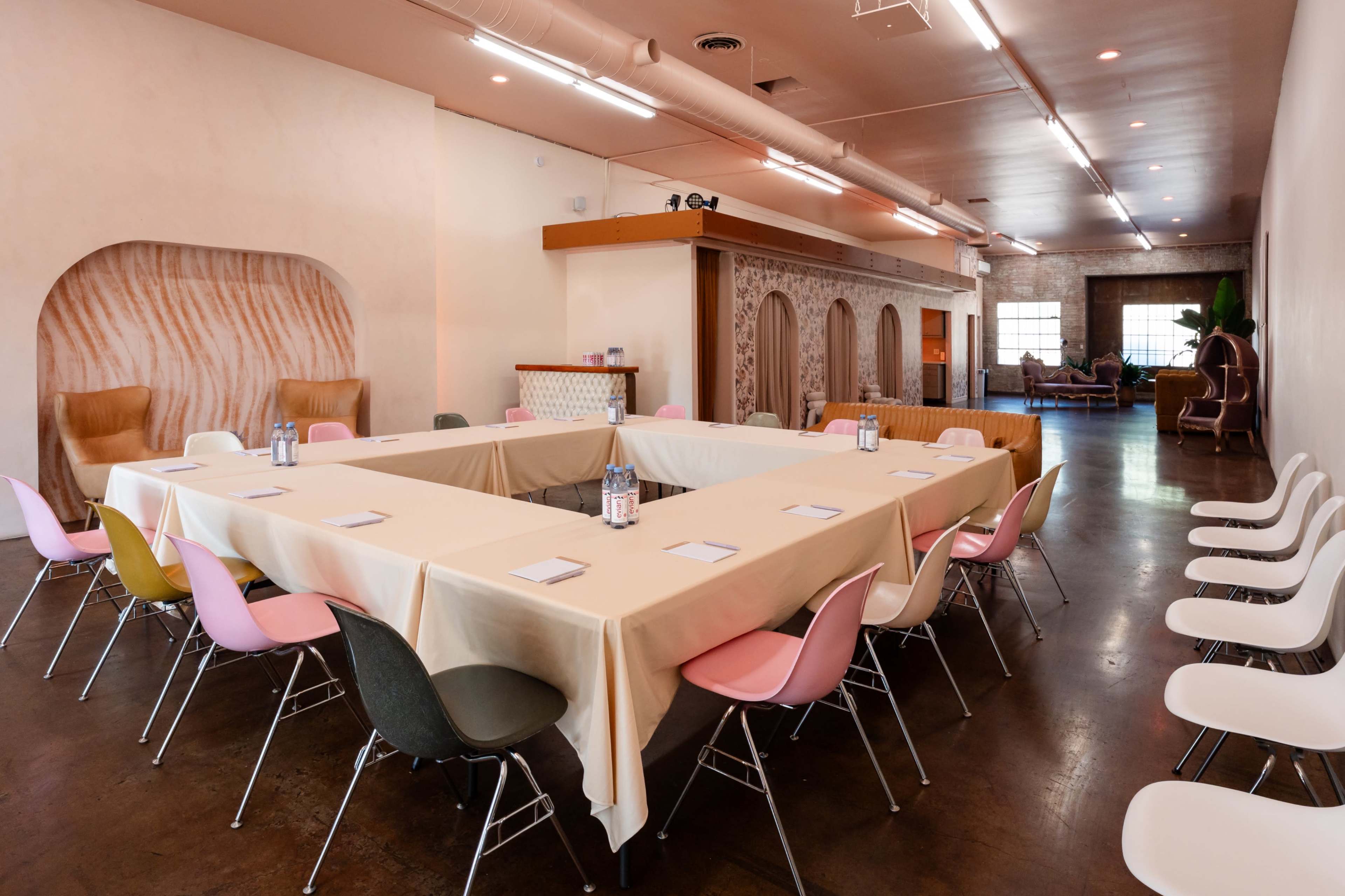 A rectangular table covered with a beige tablecloth is set in a modern meeting room, surrounded by colorful chairs and decorated with soft lighting and wall features.