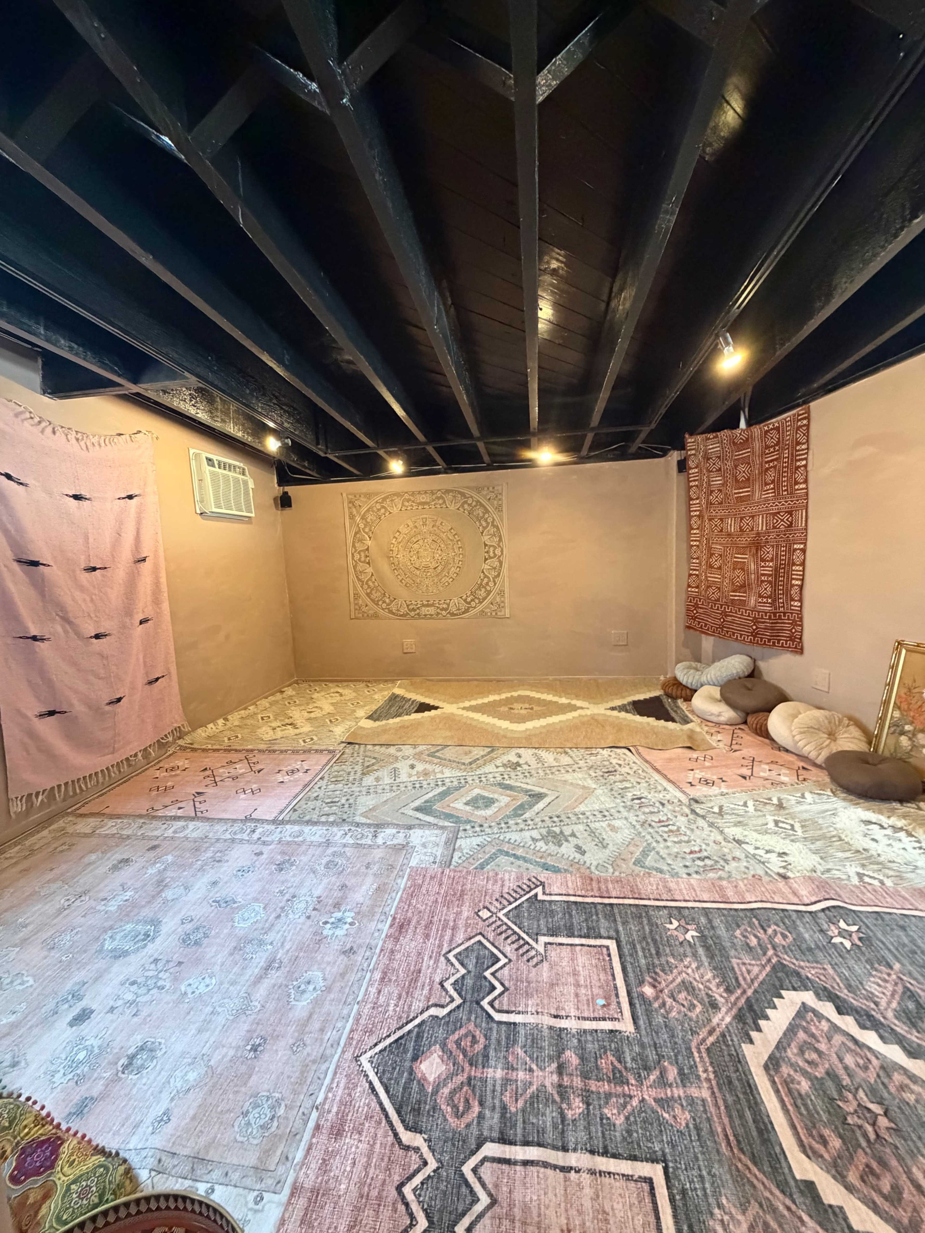 The image depicts a spacious room with decorative rugs on the floor, textured walls, and warm lighting, along with patterned wall hangings and cushions arranged in the space.