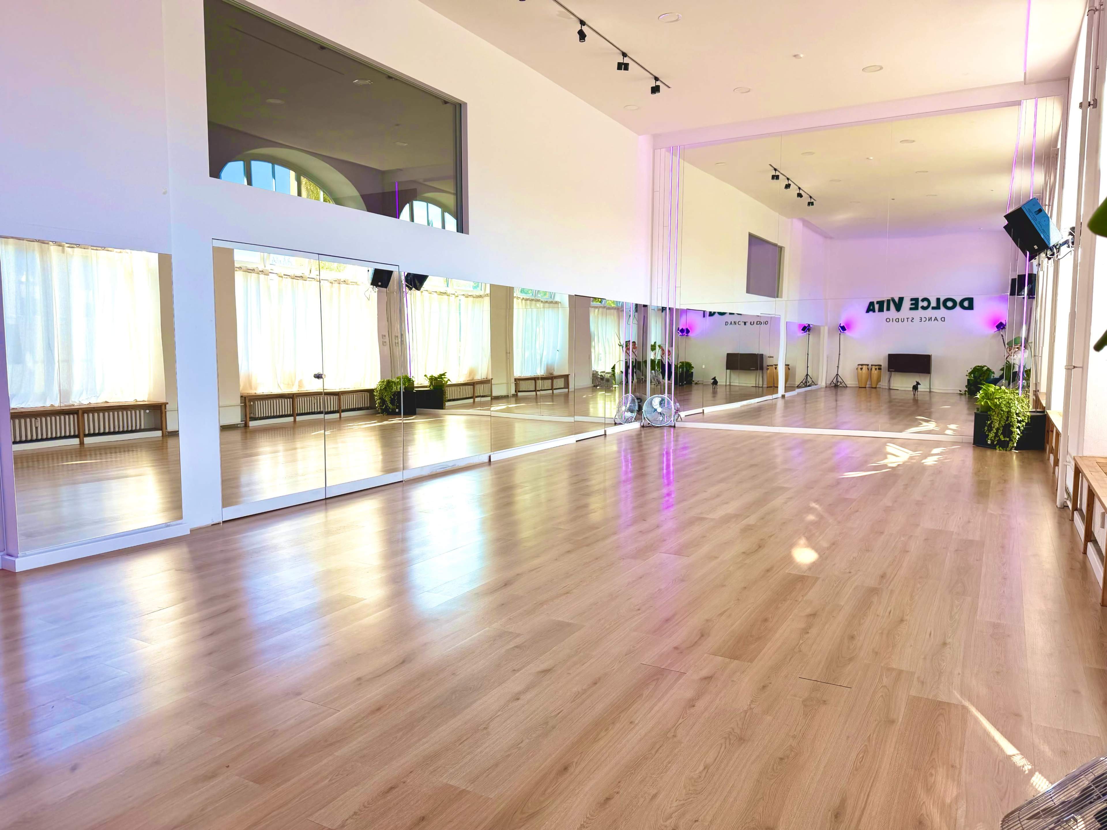 The image shows a brightly lit dance studio with large mirrors, wooden flooring, and plants, featuring purple lighting accents and large windows.