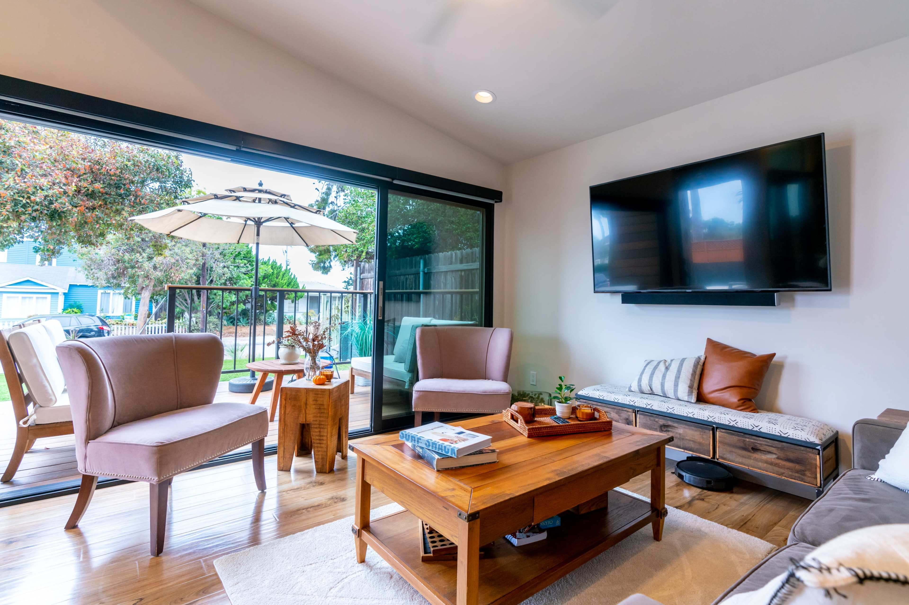 The image shows a living room with a wooden coffee table, two armchairs, a sectional sofa, and a large window opening to a patio area with an umbrella.