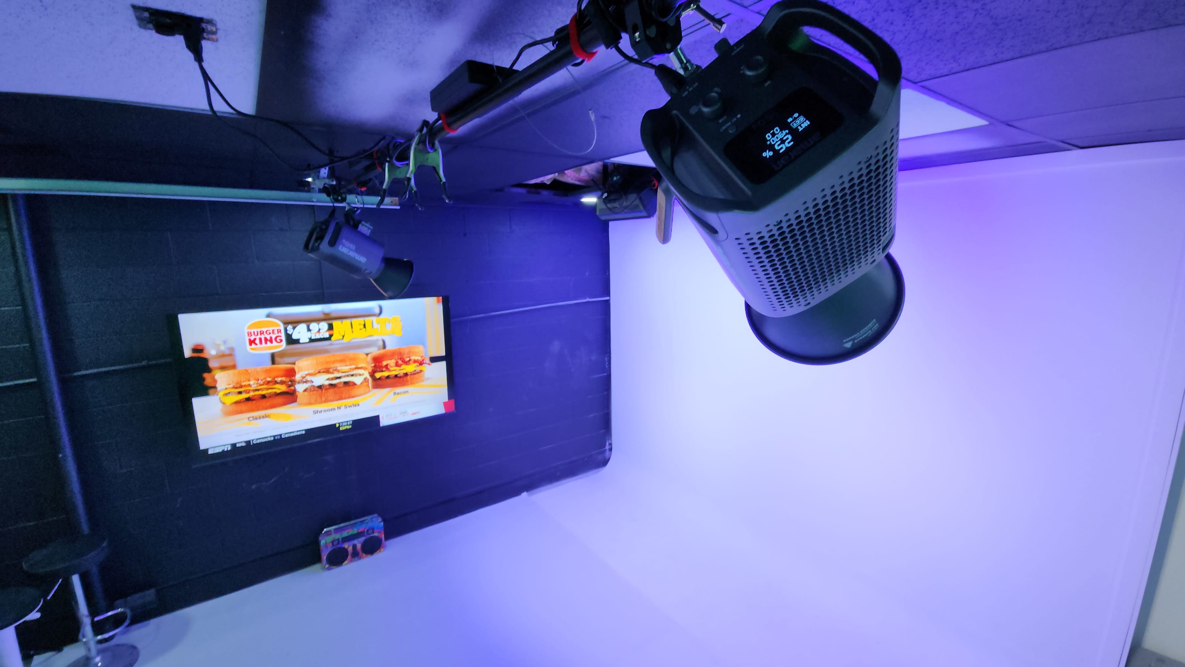 The image shows a studio or staging area with a TV displaying a fast food advertisement, a black ceiling-mounted speaker, and purple ambient lighting.