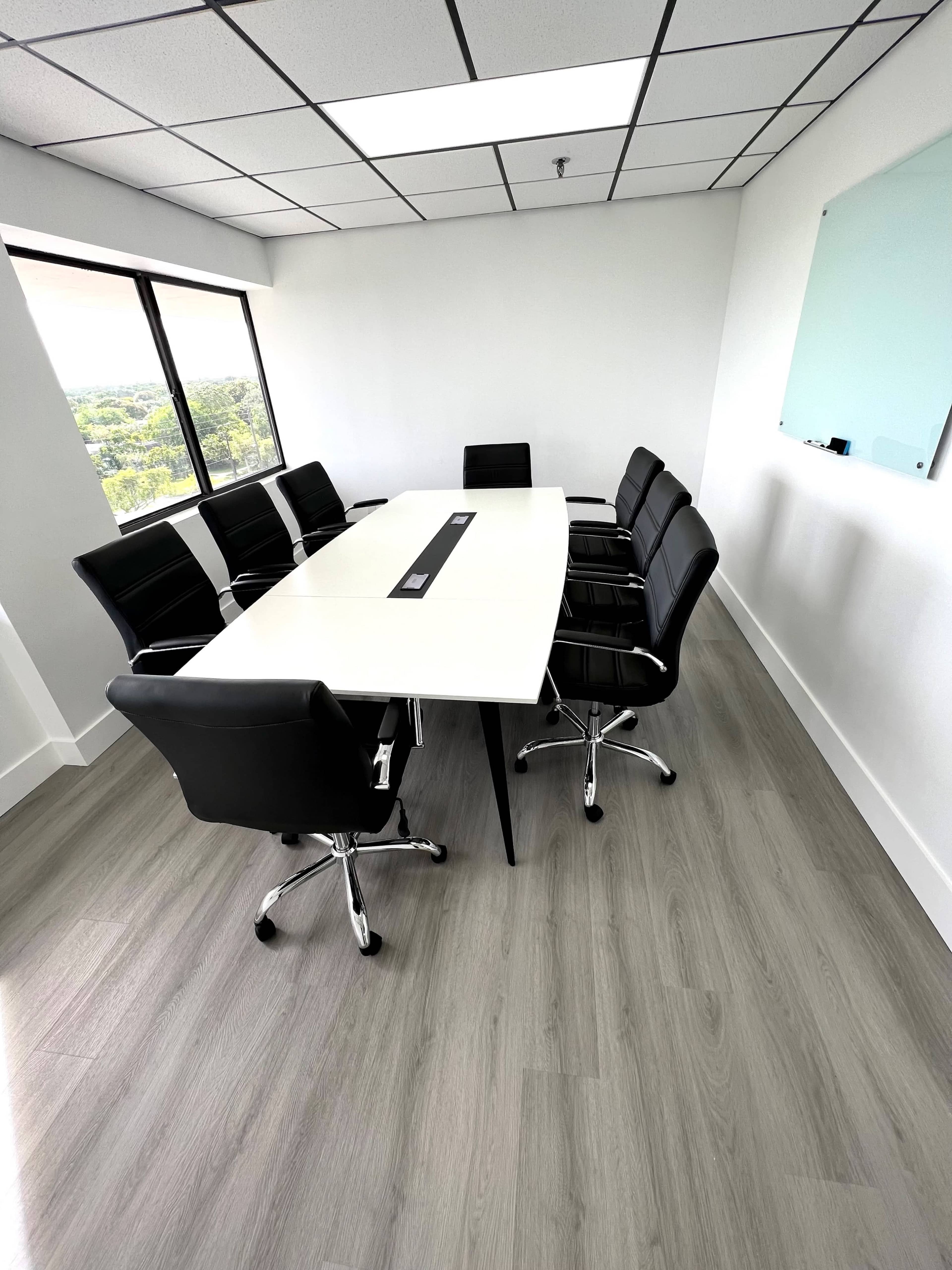 A modern conference room with a long white table surrounded by black chairs and large windows overlooking a green landscape.