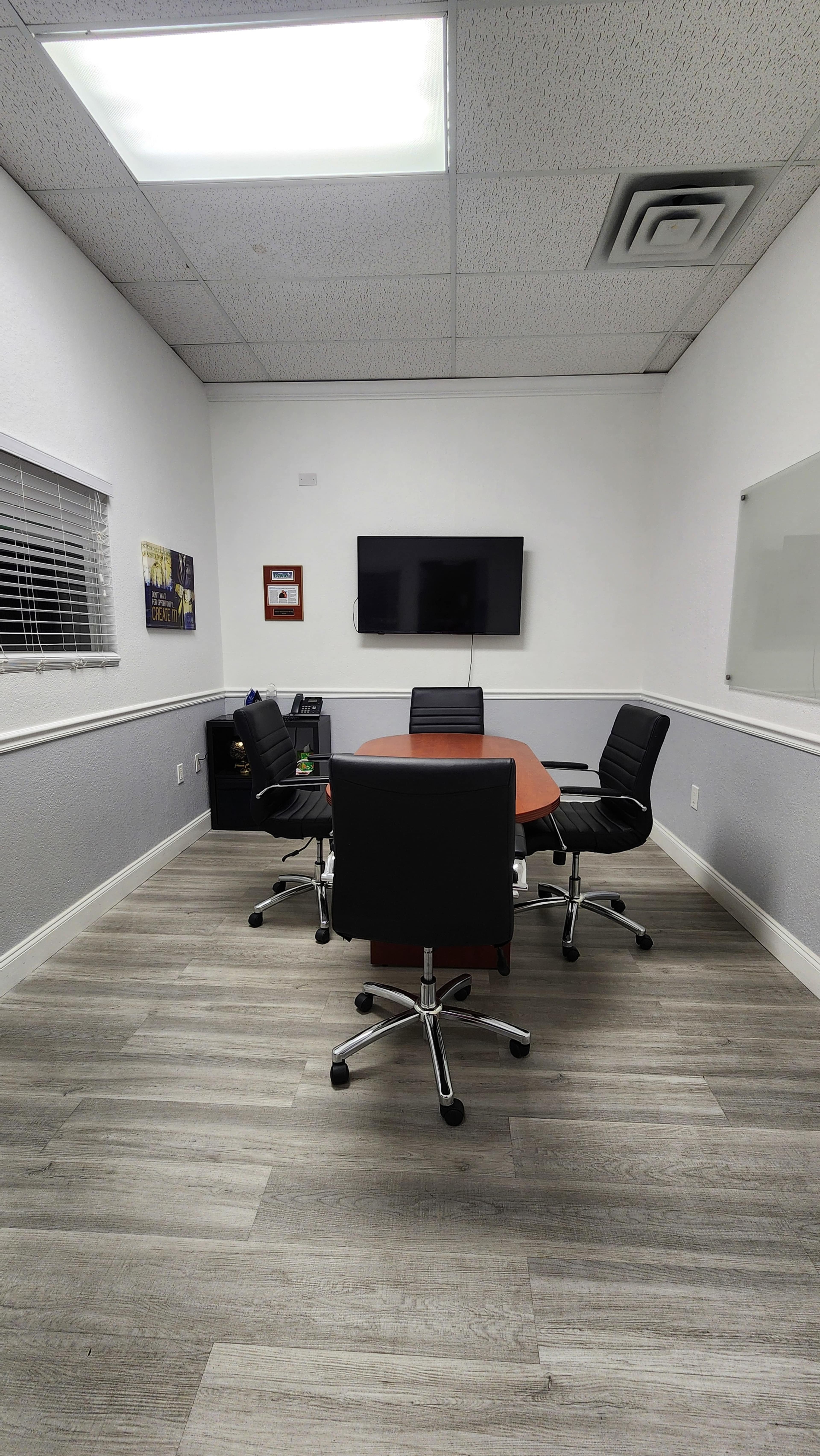 A small conference room features a round wooden table surrounded by black rolling chairs, with a television mounted on the white wall.