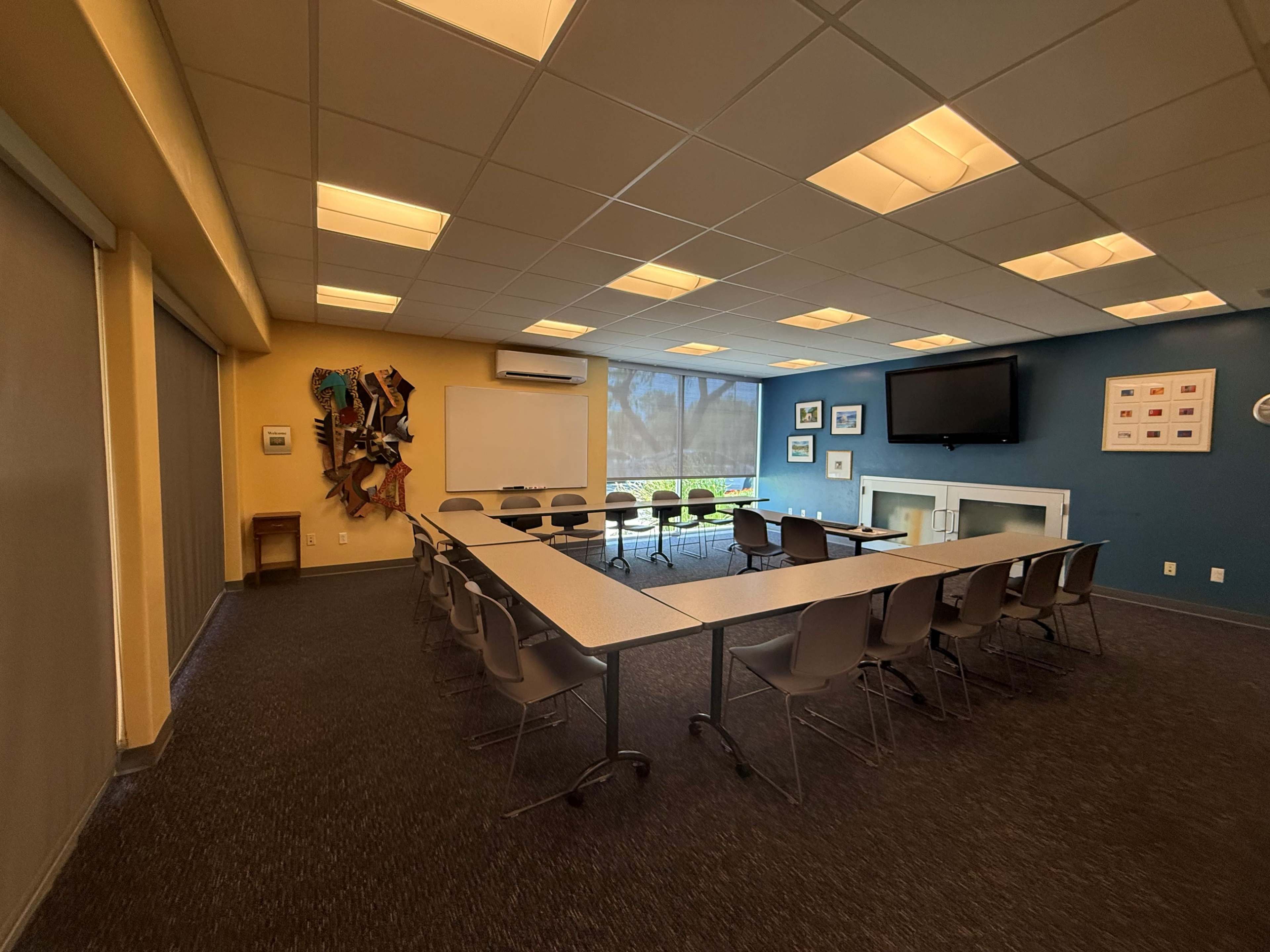 The image shows a rectangular meeting room with a long table surrounded by chairs, and large windows allowing natural light to enter.