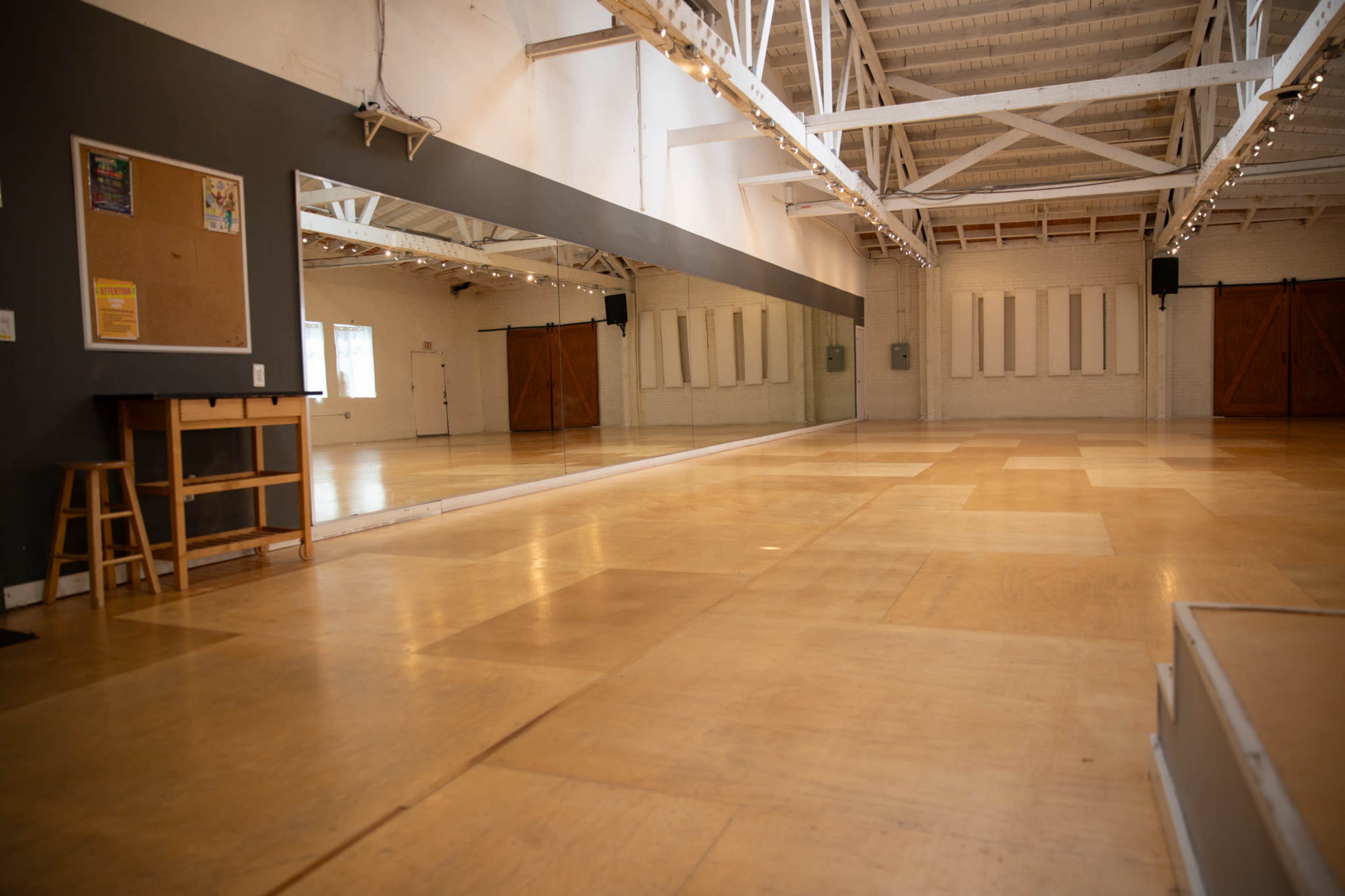 The image shows a spacious dance studio with wooden floors, mirrors along one wall, and a set of double doors at the far end.
