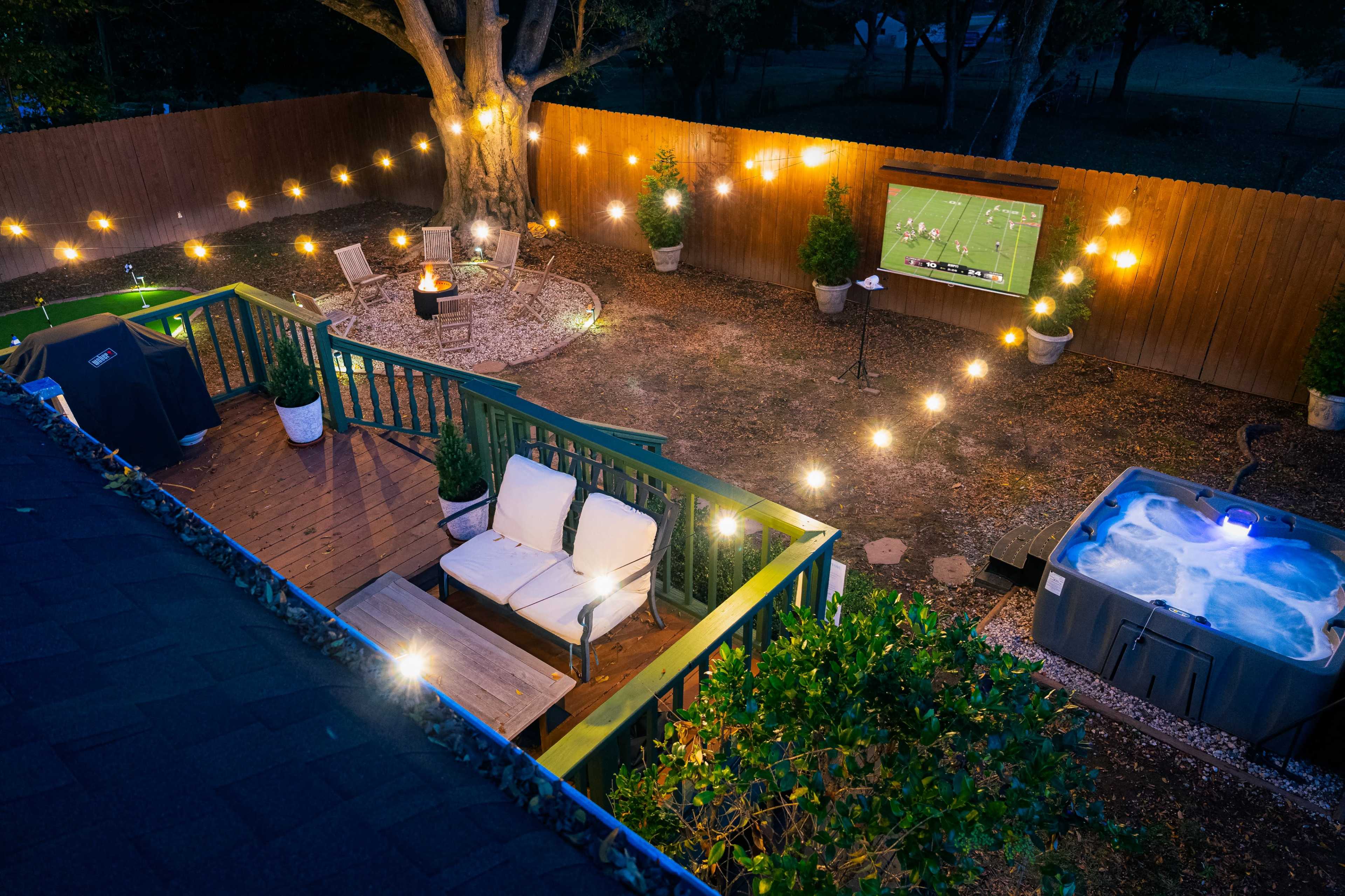The image shows a well-lit backyard with a seating area, a hot tub, and a screen displaying a sports game, surrounded by decorative plants and a wooden fence.
