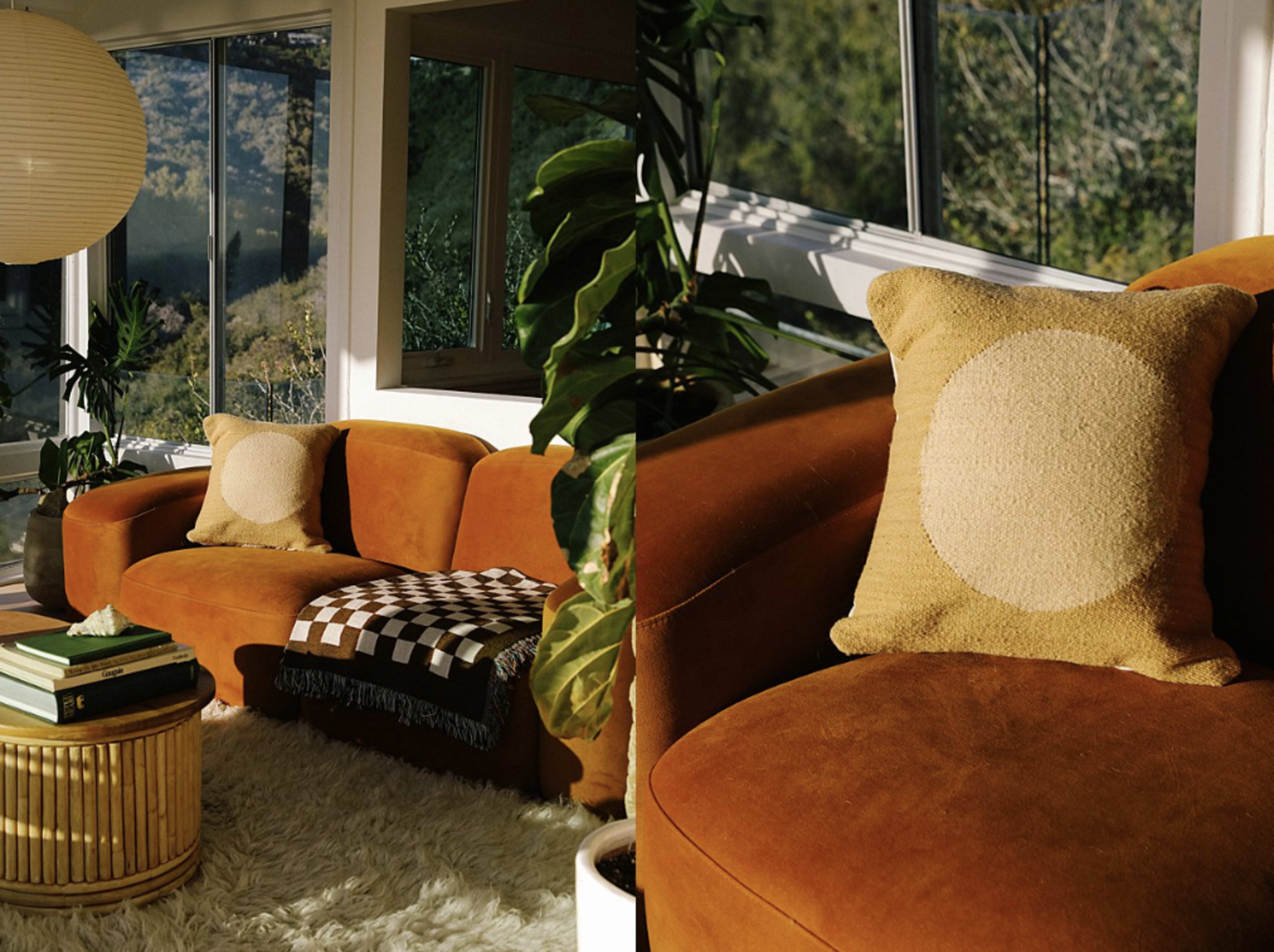 A curved orange sofa with a decorative beige pillow is positioned in a sunlit room featuring large windows and green plants.