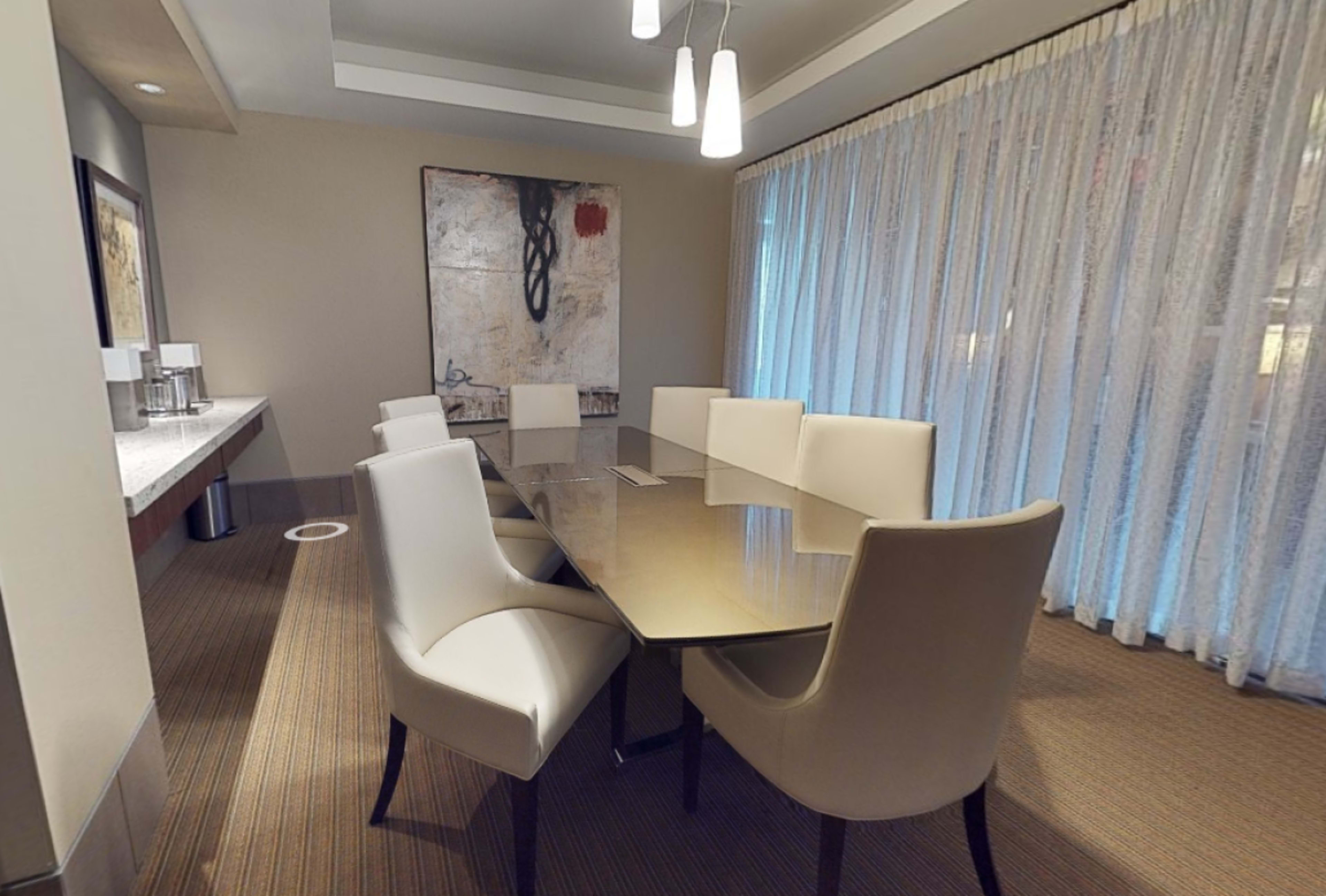 A conference room features a rectangular glass table surrounded by eight white upholstered chairs, with a large artwork on the wall and sheer curtains covering a window.