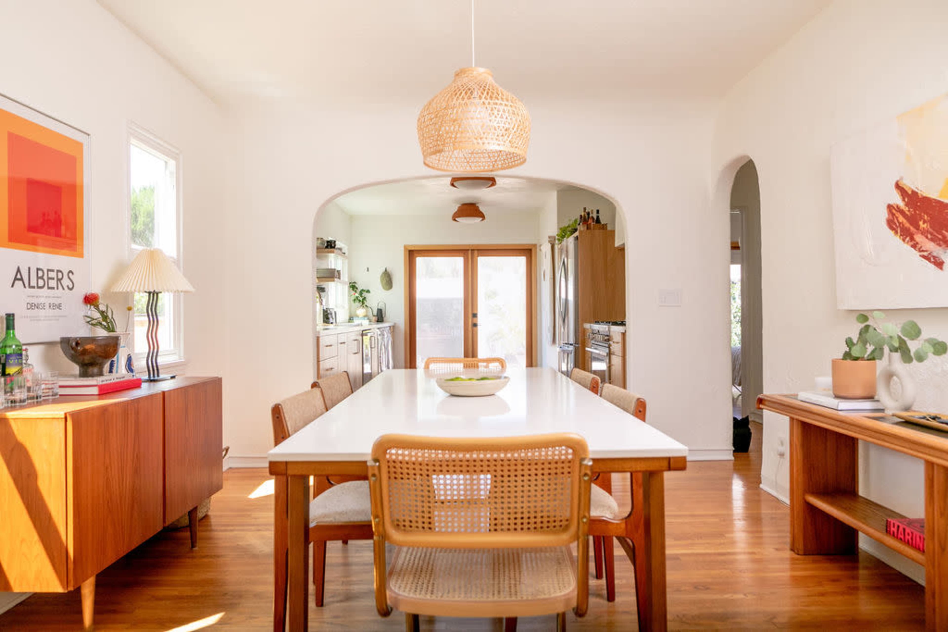 A spacious dining area features a white table surrounded by wooden chairs, illuminated by a woven pendant light, and leading to an arched doorway.