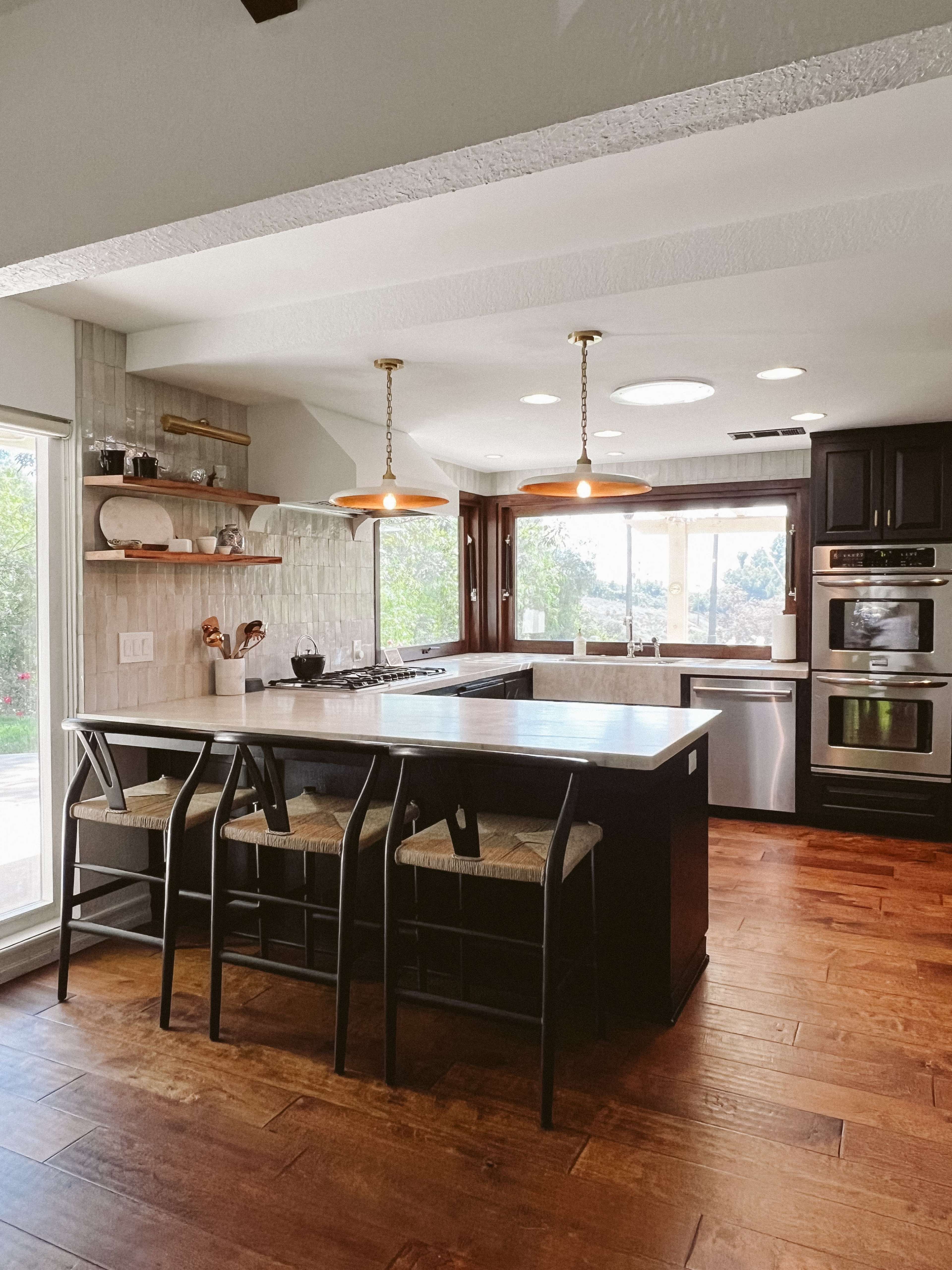 A modern kitchen features a central island with bar seating, stainless steel appliances, and large windows providing natural light.