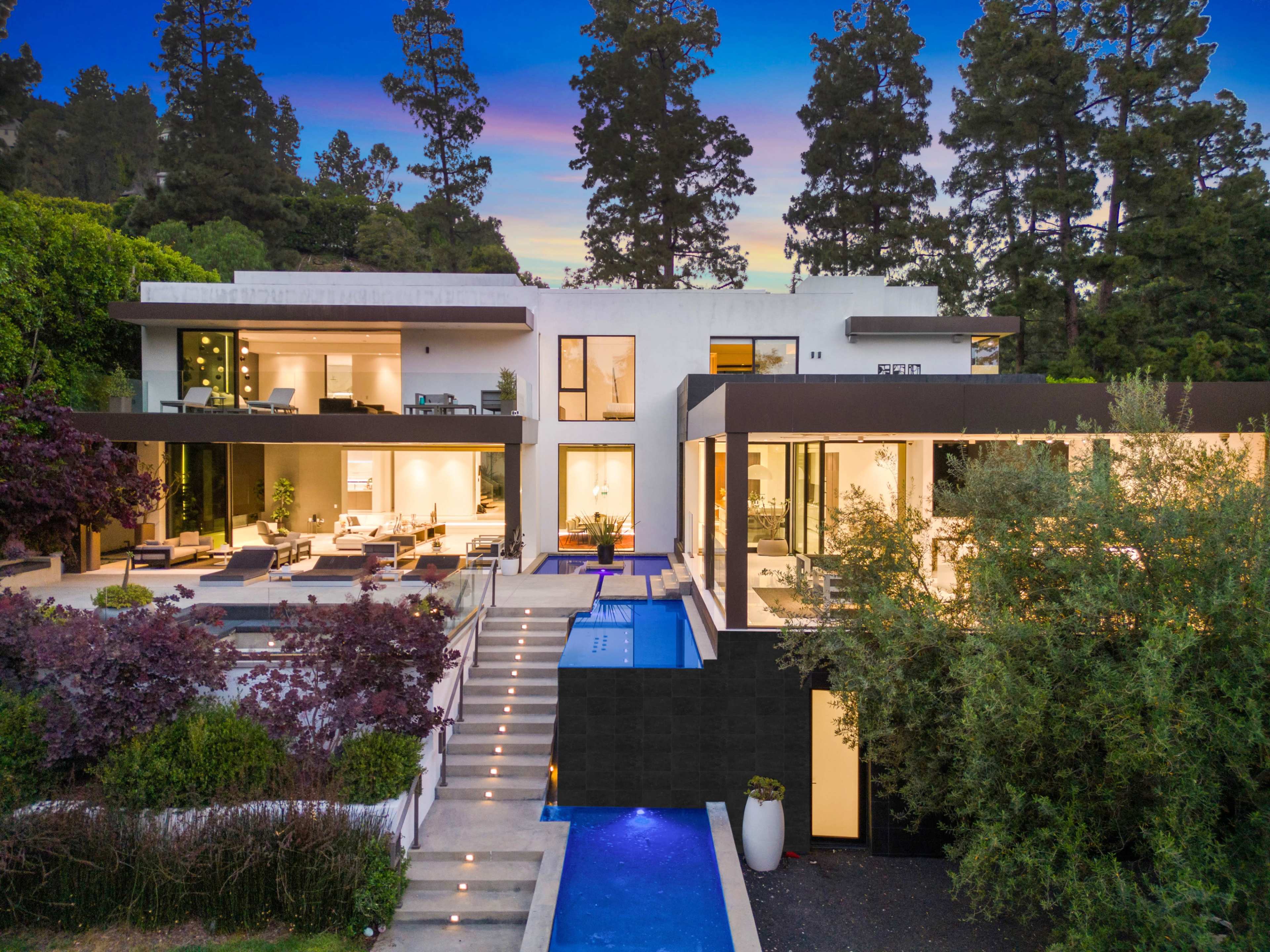 The image shows a modern two-story house surrounded by trees, featuring a pool and a landscaped yard with various plants.